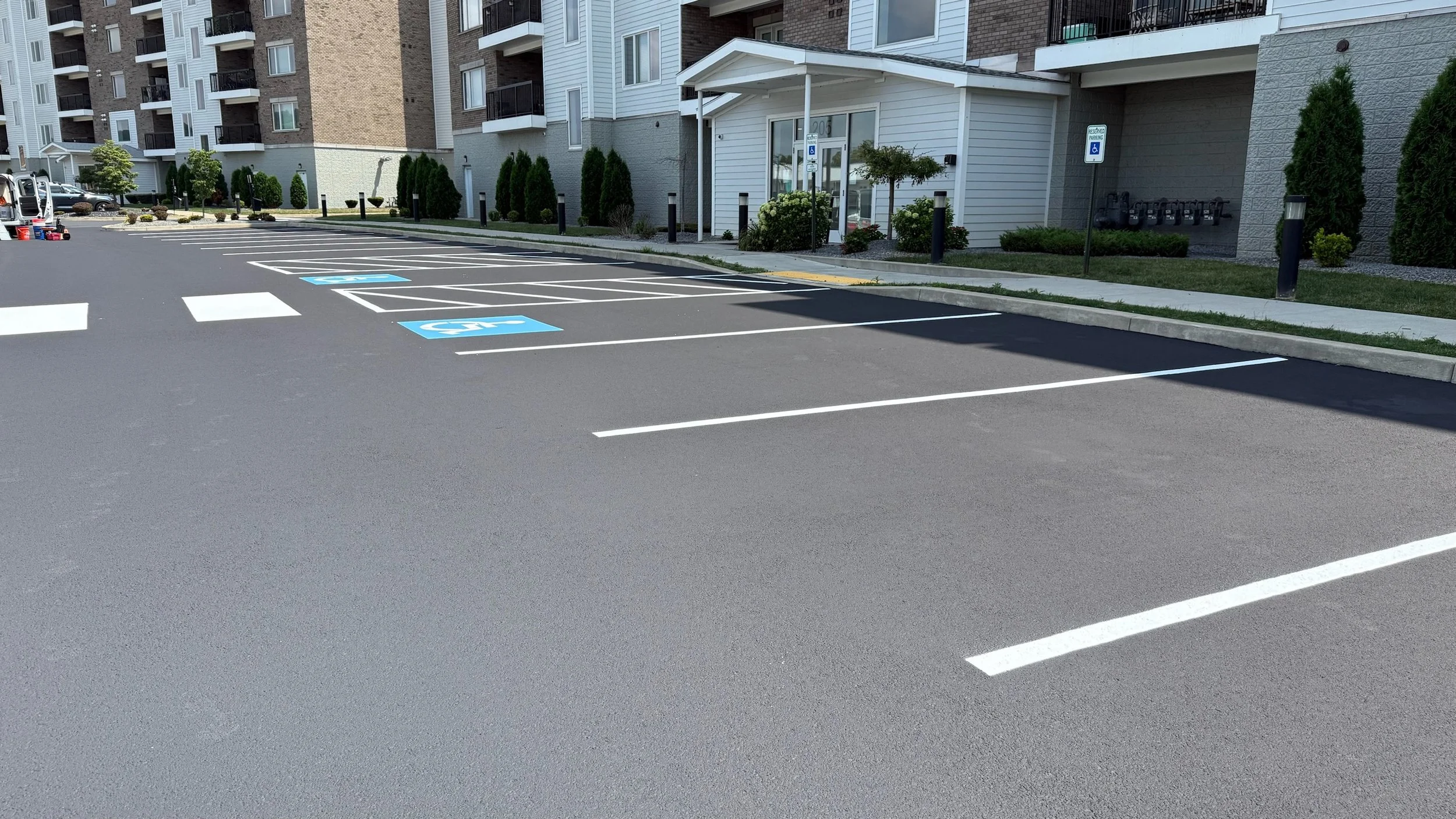 Empty parking lot with designated handicap spots marked with blue signs and white wheelchair symbols, adjacent to an apartment building with landscaped bushes and trees.