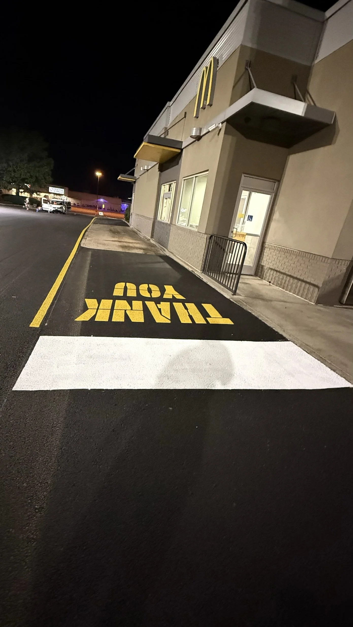 Nighttime view of a McDonald's drive-thru lane with yellow painted words reading "THANK YOU" on the black asphalt and a white pedestrian crosswalk. The building has large windows and is lit from inside. There are parked cars in the distance.