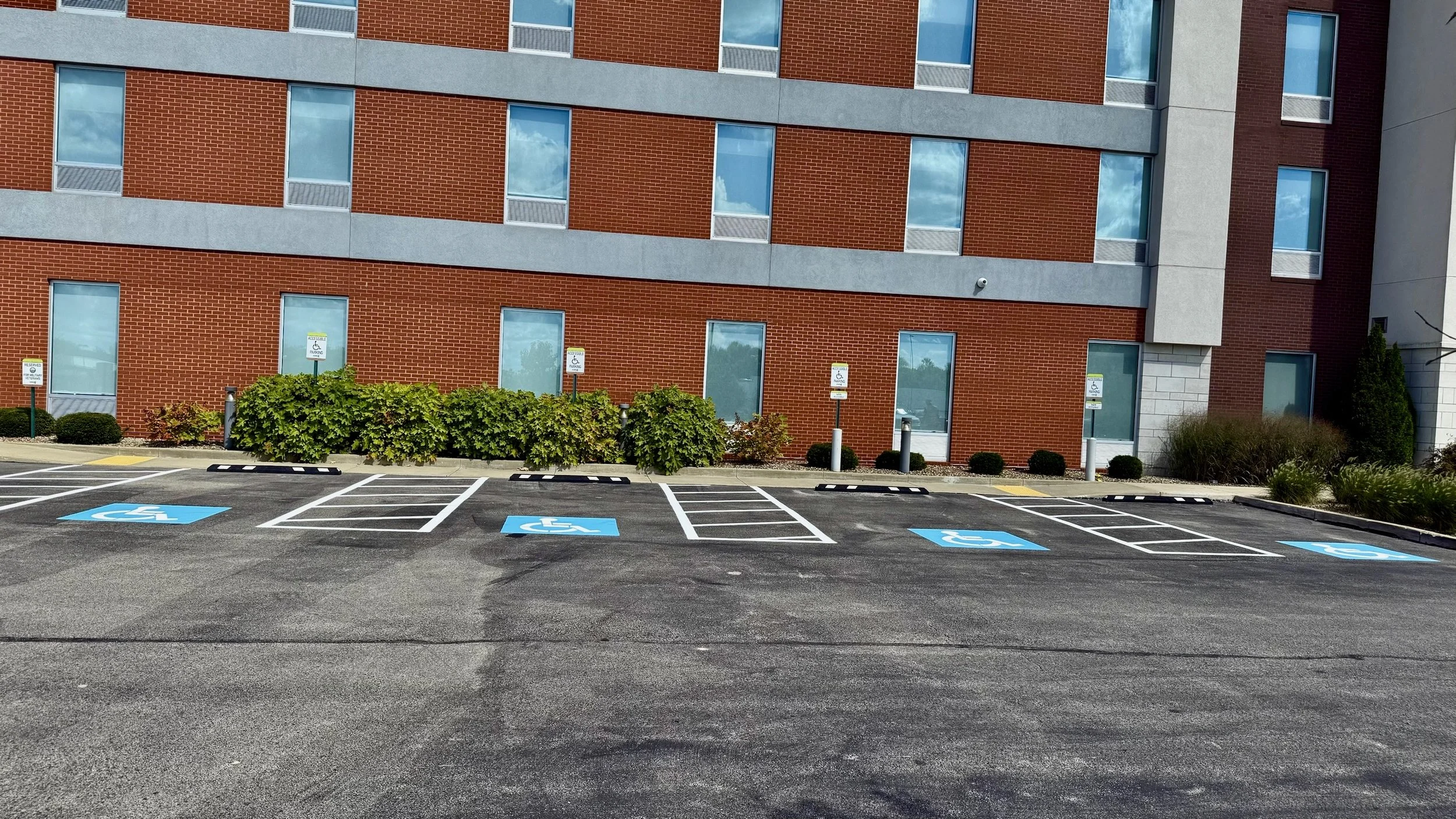 Empty parking lot with designated accessible parking spaces marked with blue and white signs and pavement symbols, in front of a multi-story brick building with several windows and landscaped bushes.