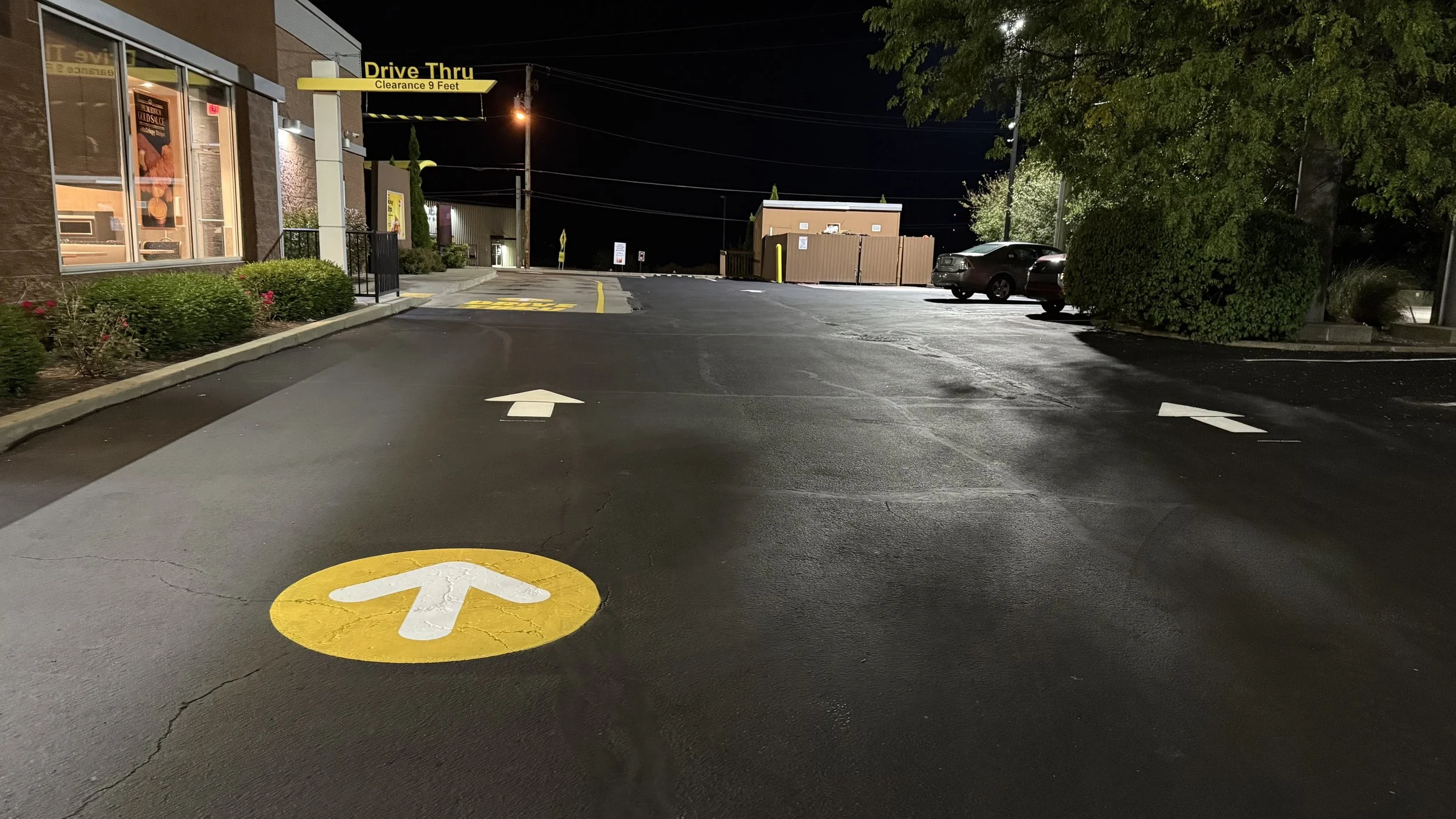 Nighttime view of a McDonald's restaurant drive-thru with a yellow and white arrow on the pavement indicating the drive-thru lane. The area is illuminated by streetlights and the restaurant's exterior lighting, with parked cars and lush green bushes