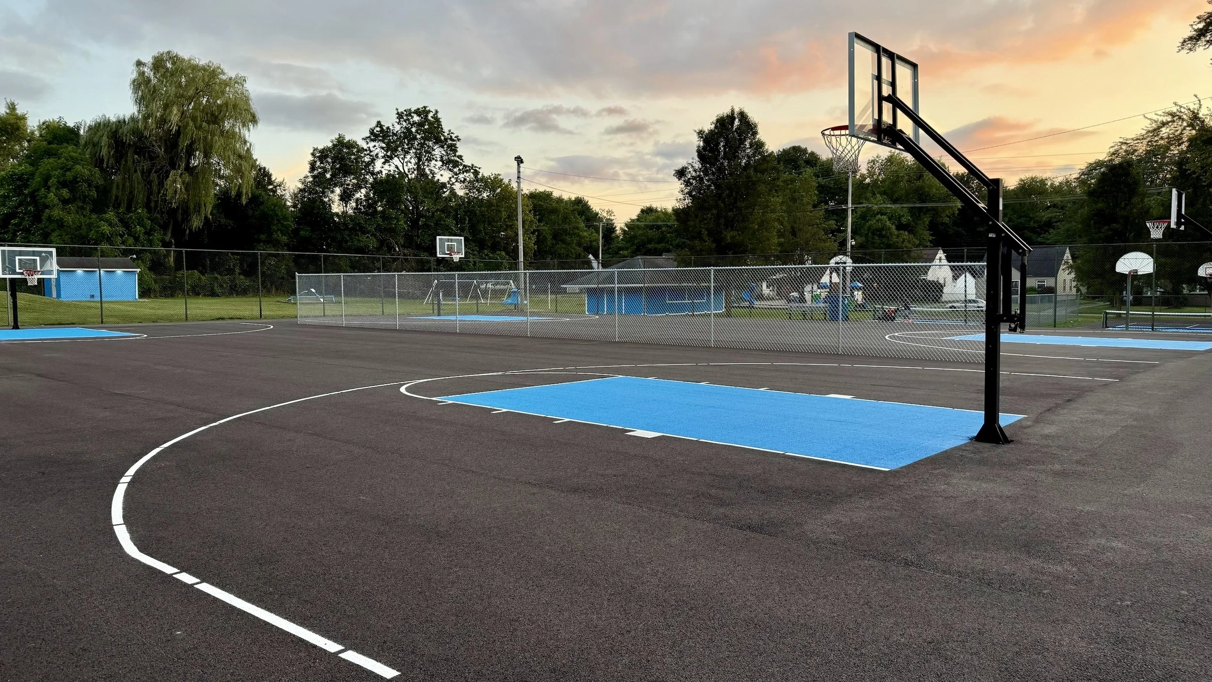 An empty outdoor basketball court at sunset, with blue painted key areas and a single basketball hoop.