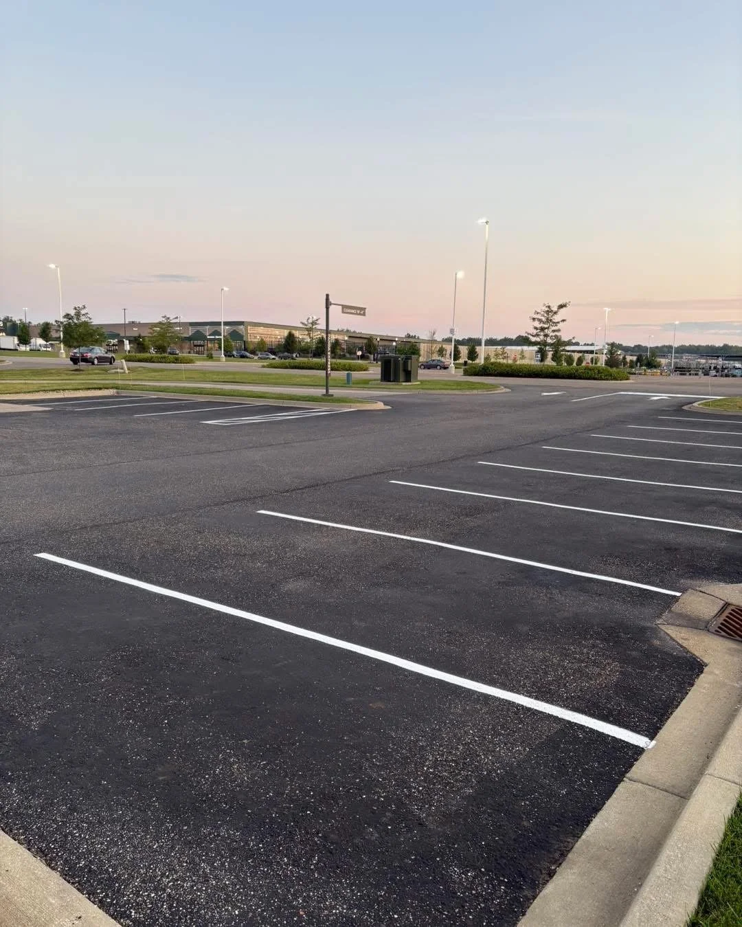 Empty parking lot at dusk with a few streetlights, a shopping center in the background, and trees.