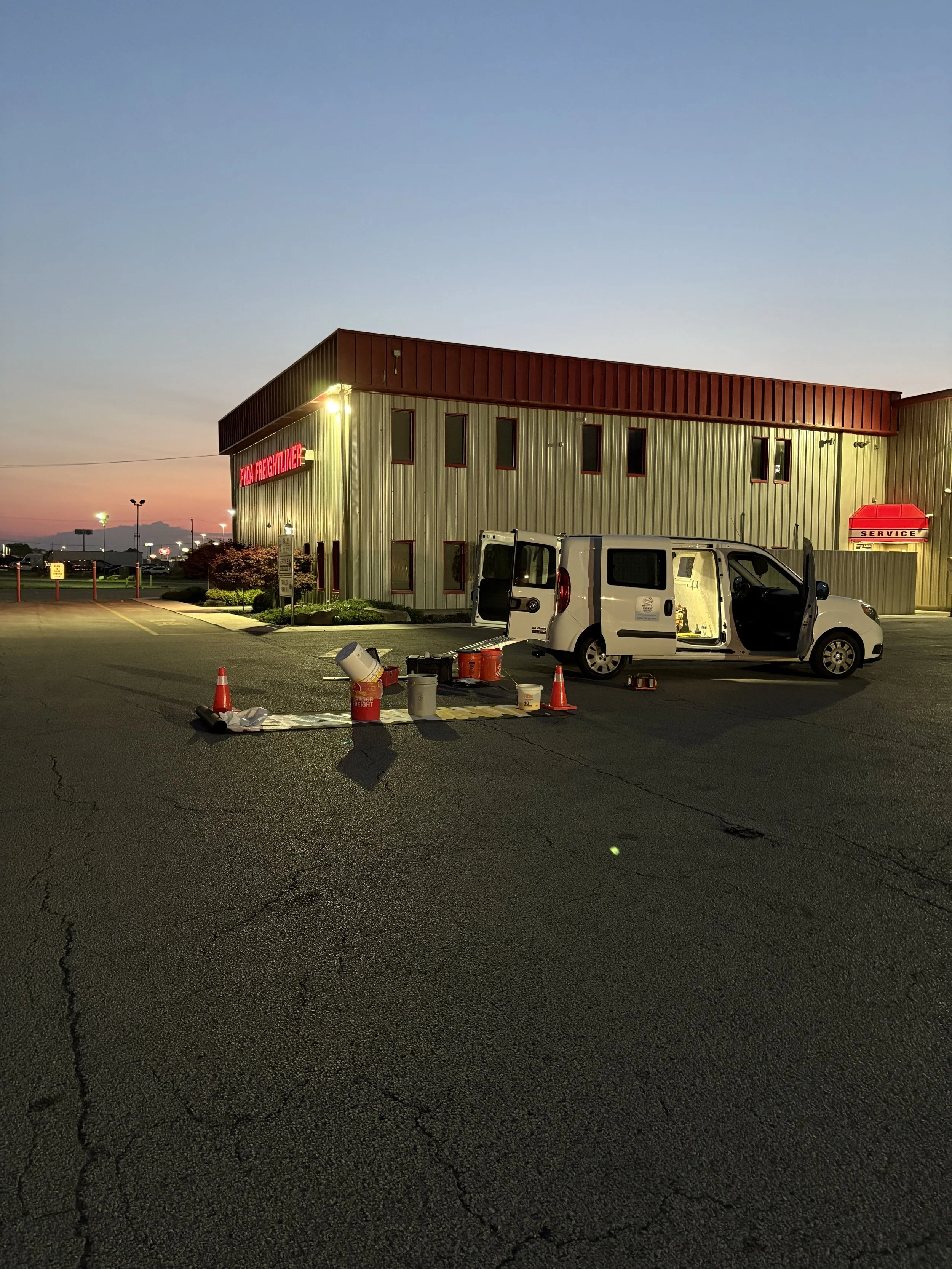 Parking lot outside a building at dusk with a white work van parked, open doors, and various supplies, including cones, buckets, and tools, on the ground.