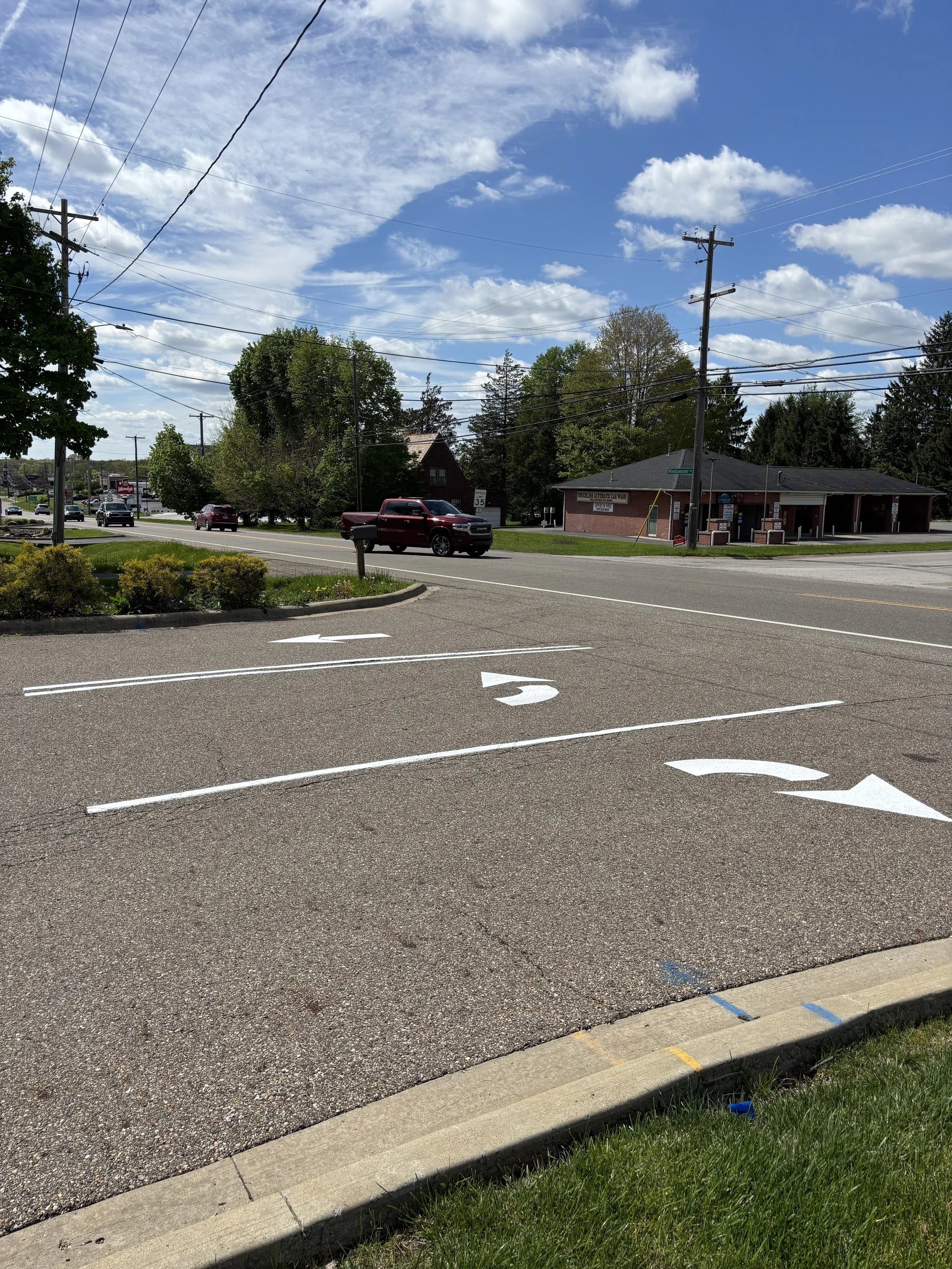 Empty parking lot with white arrows painted on the asphalt indicating direction, surrounded by green plants, trees, a brick building, and a blue sky with clouds.
