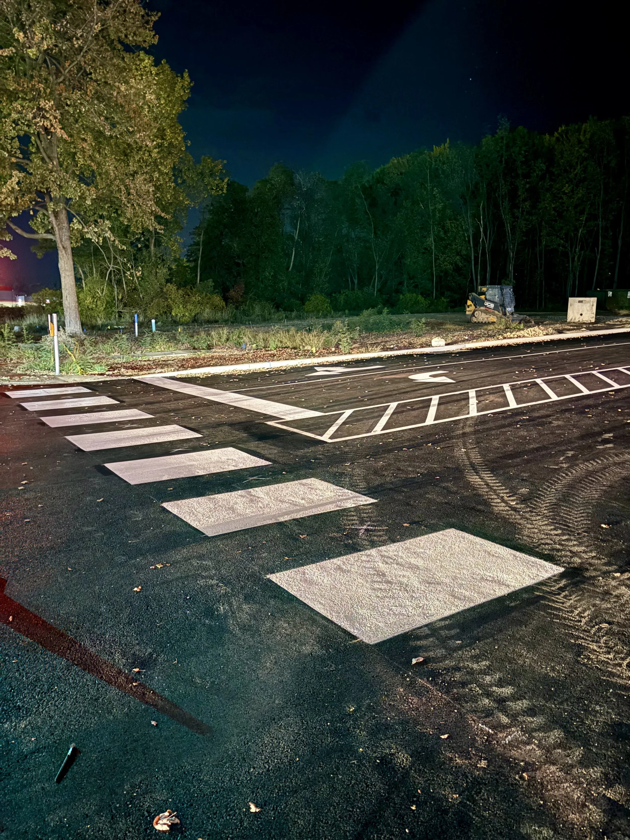 Nighttime view of a newly paved road with freshly painted pedestrian crosswalk and parking lot markings, adjacent to wooded area with trees and construction equipment in the background.