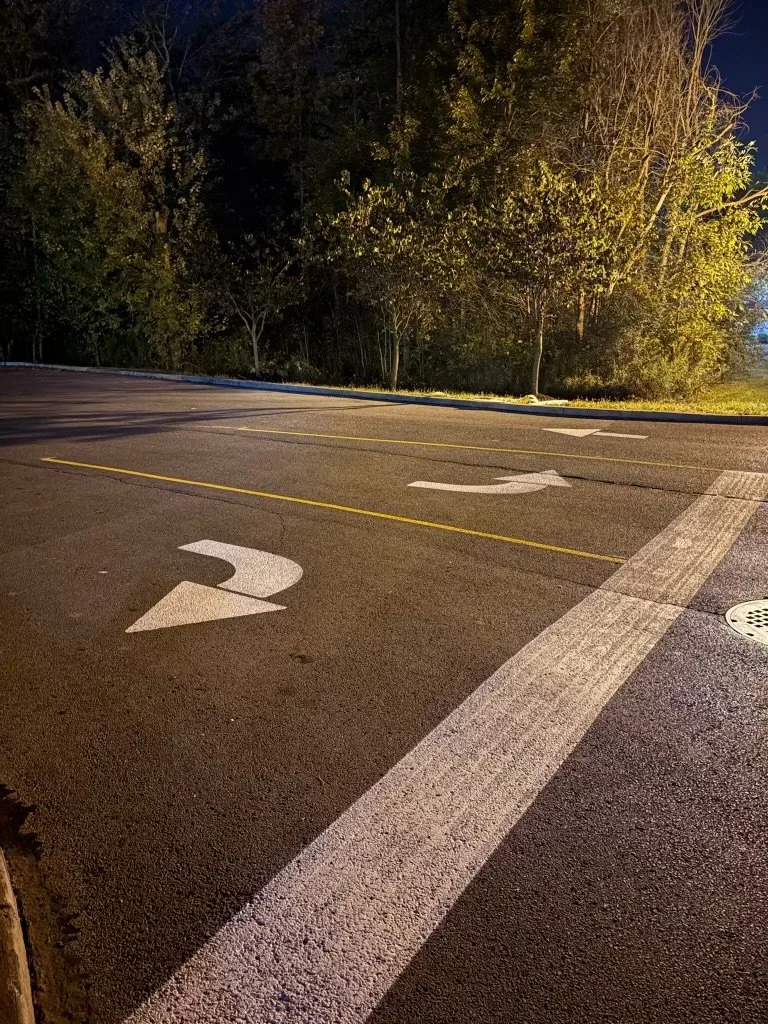 A parking lot at night with lane markings and directional arrows, empty and illuminated by streetlights, with trees in the background.