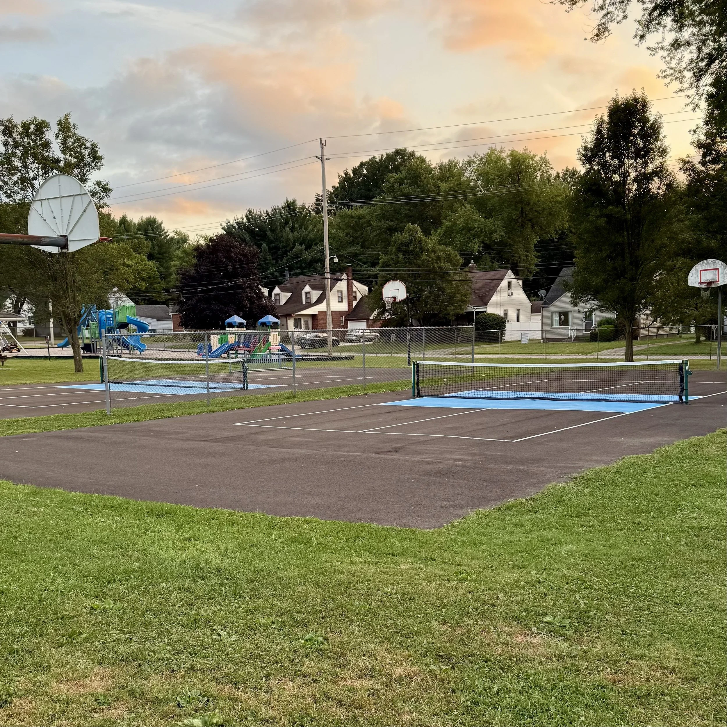 A playground with basketball courts and tennis courts, surrounded by trees and houses during sunset.