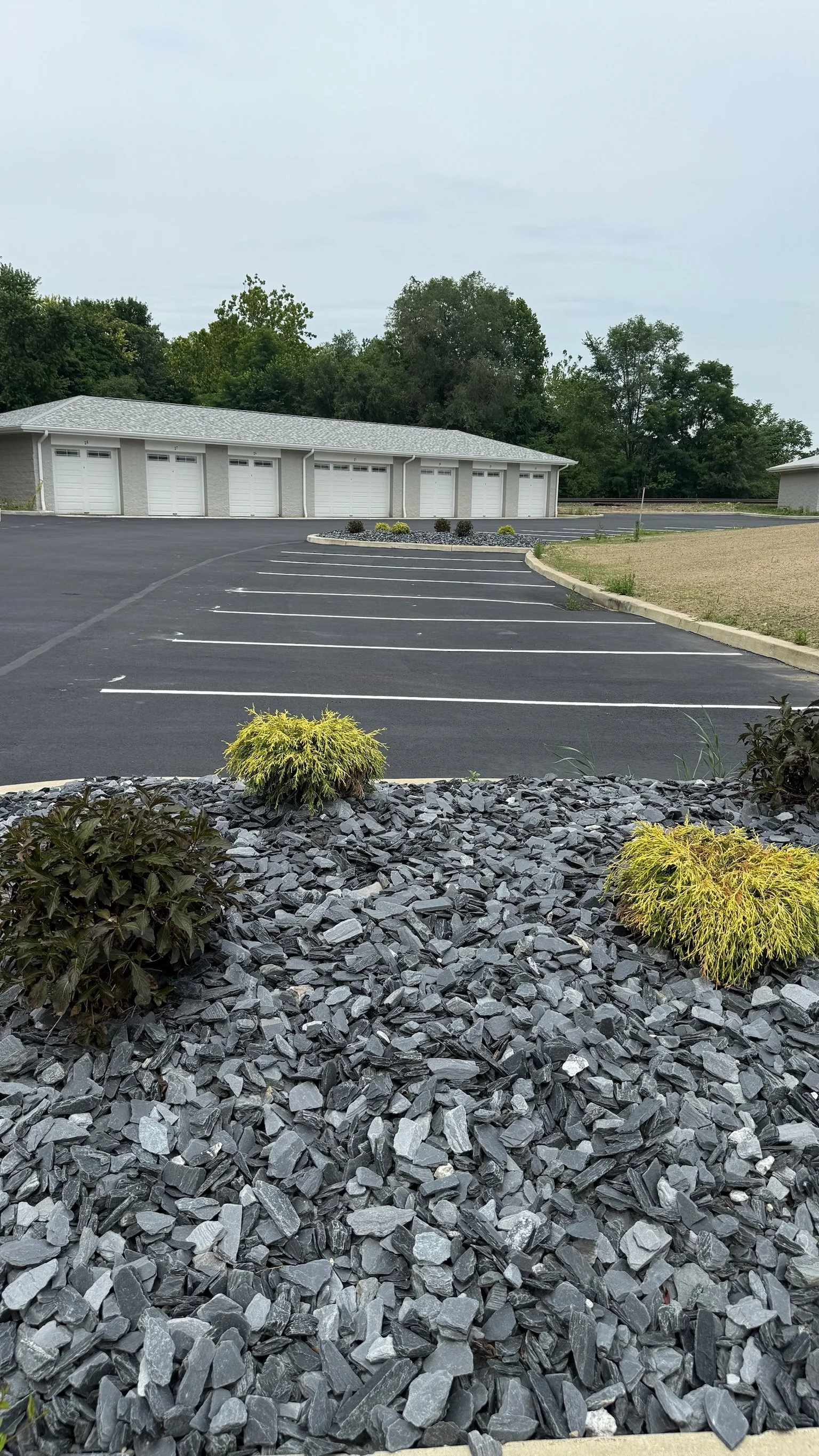 Empty parking lot with paved and gravel areas, bushes, and a building with multiple garage doors in the background.