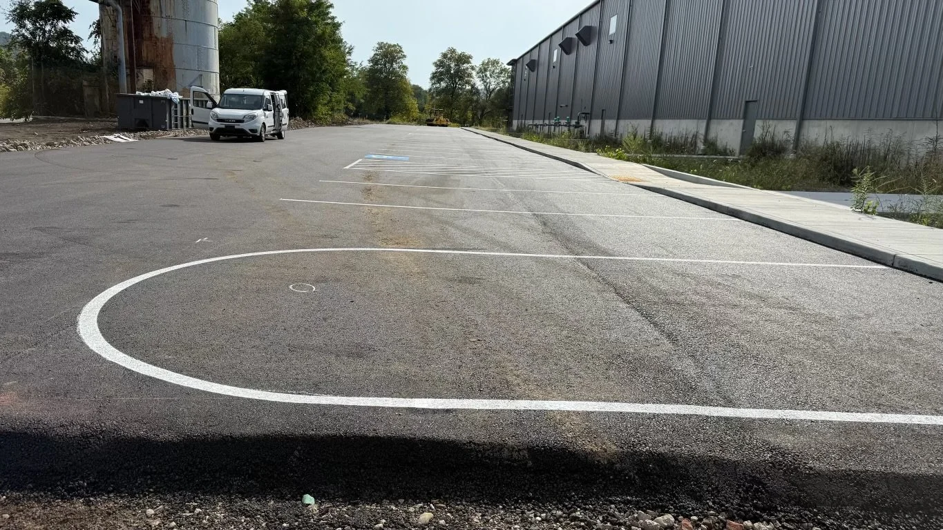 Empty outdoor parking lot with a small section of painted basketball court visible in the foreground, a silver van parked near a trash bin on the left, trees, and a large industrial building on the right.