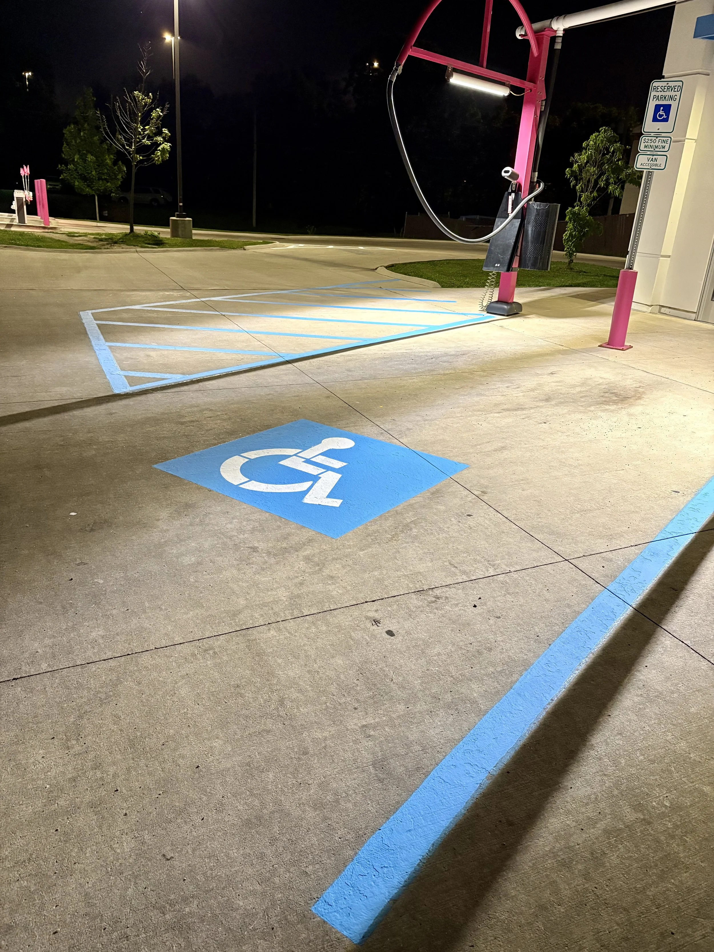 Empty wheelchair accessible parking space with blue painted handicap symbol on concrete, marked by blue lines, at night outside a building with a pink lift device nearby.