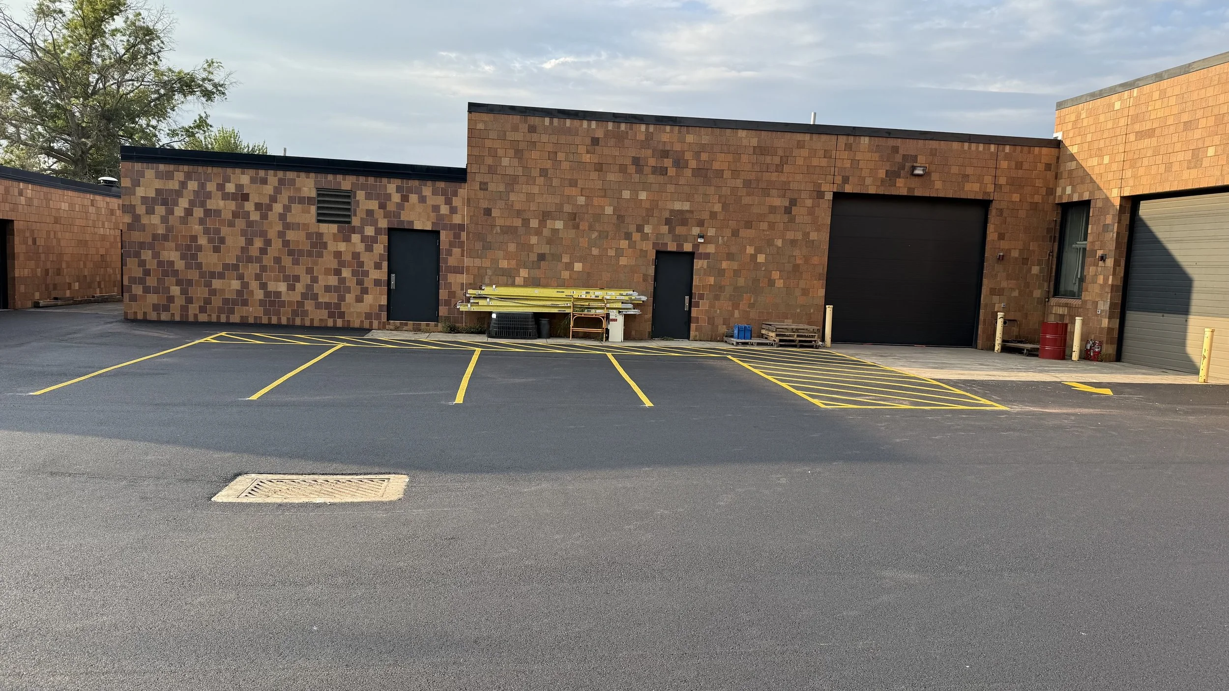 Empty parking lot with yellow striped parking spaces in front of a brick building with three doors and various equipment.
