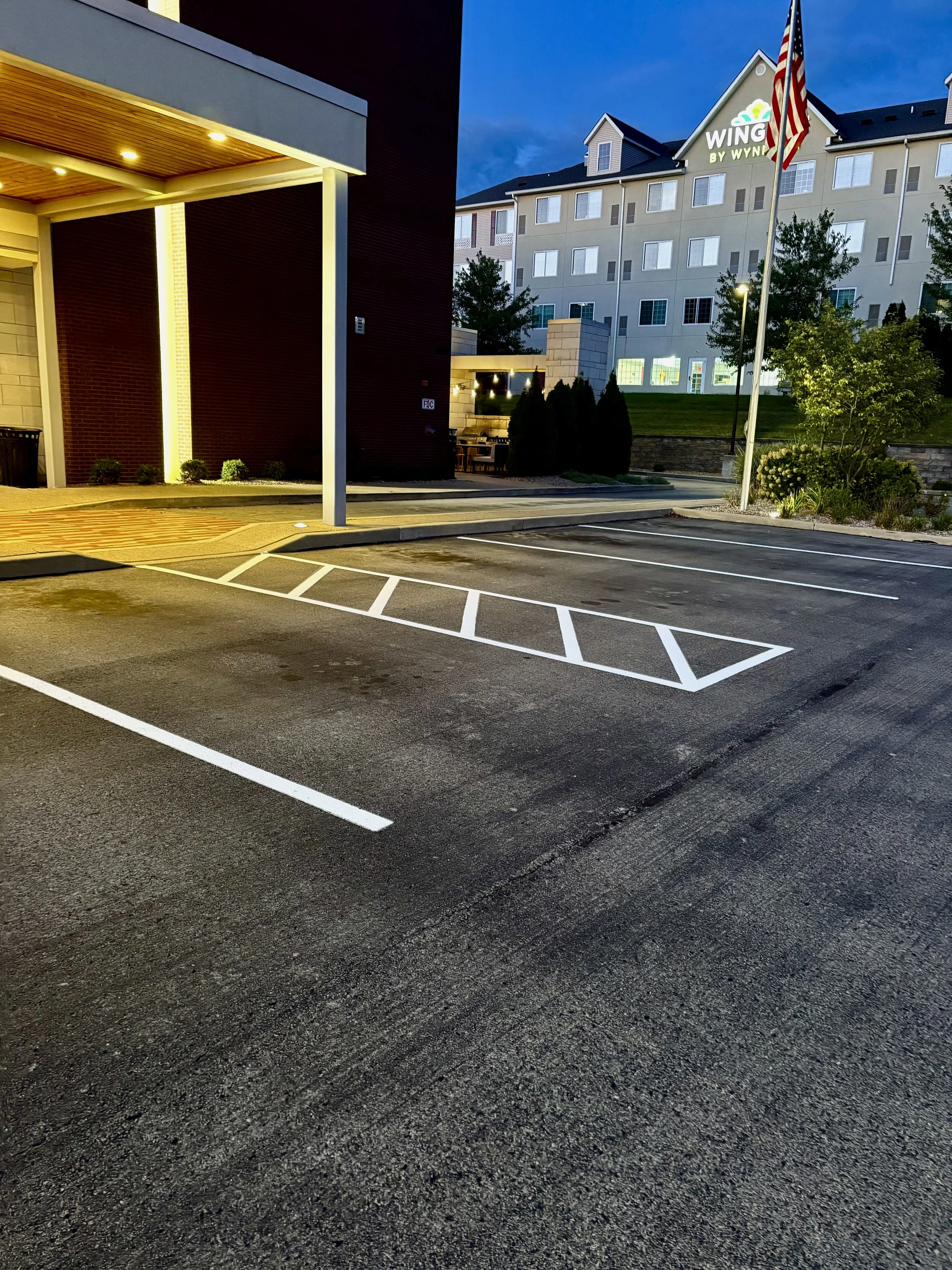 Empty parking lot at dusk with a building and trees in the background, American flag flying, and a sign reading 'Wing By Wyn' on a hill.