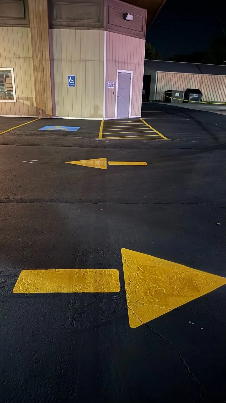 Parking lot at night with yellow directional arrows and lines, a handicapped parking sign, a building with a door, and dumpsters in the background.