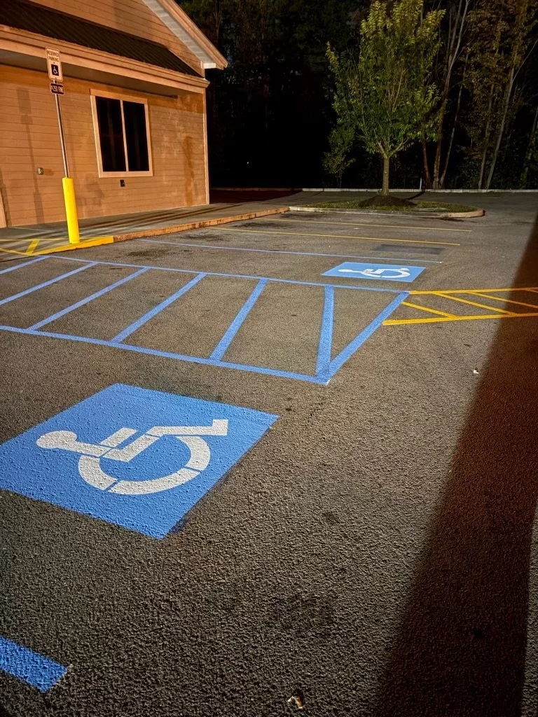 Empty parking lot at night with marked handicap parking spaces and a tree near a wooden building.