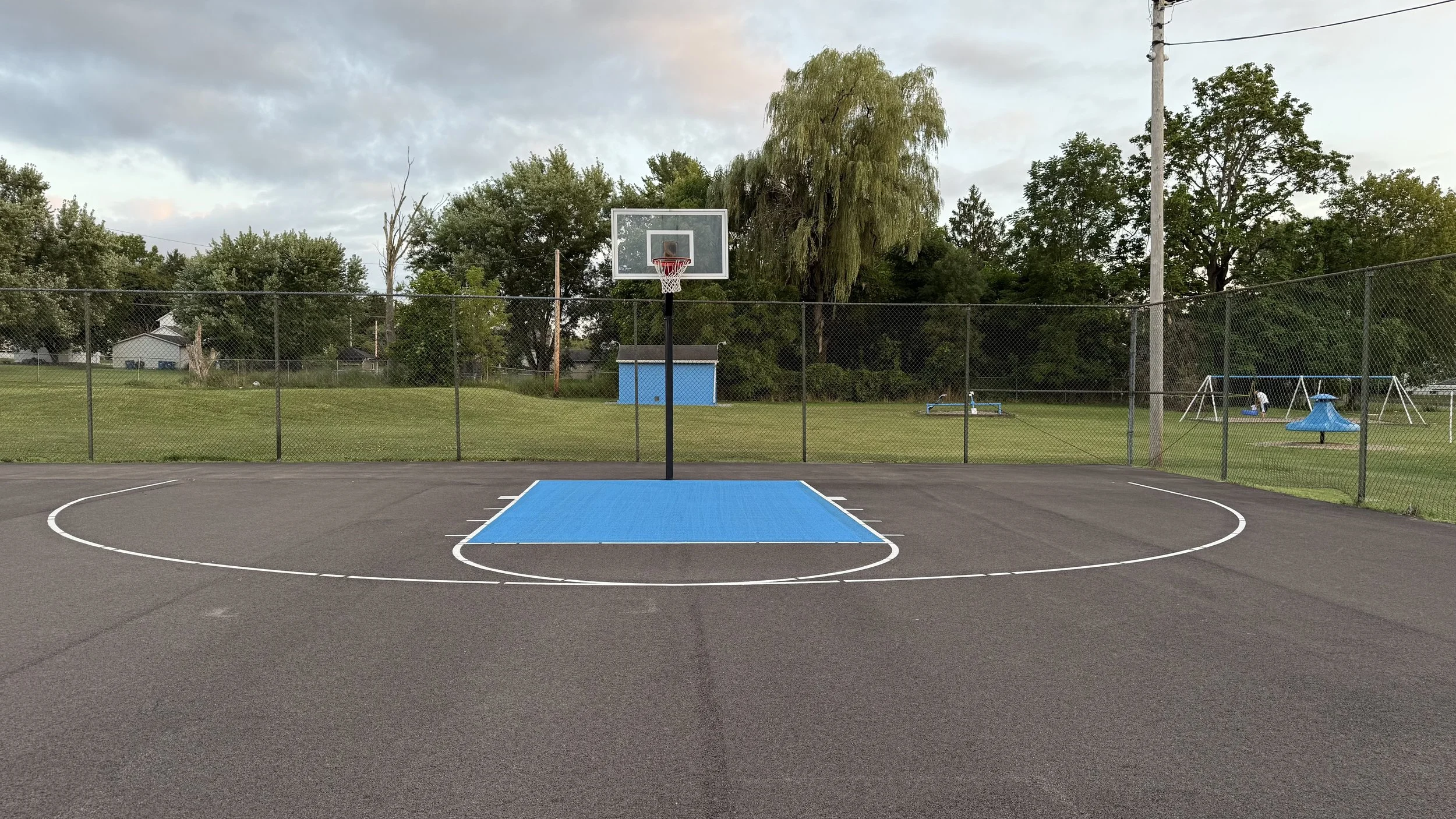 Empty outdoor basketball court with a hoop and backboard, surrounded by a chain-link fence, with a playground and trees in the background under a cloudy sky.