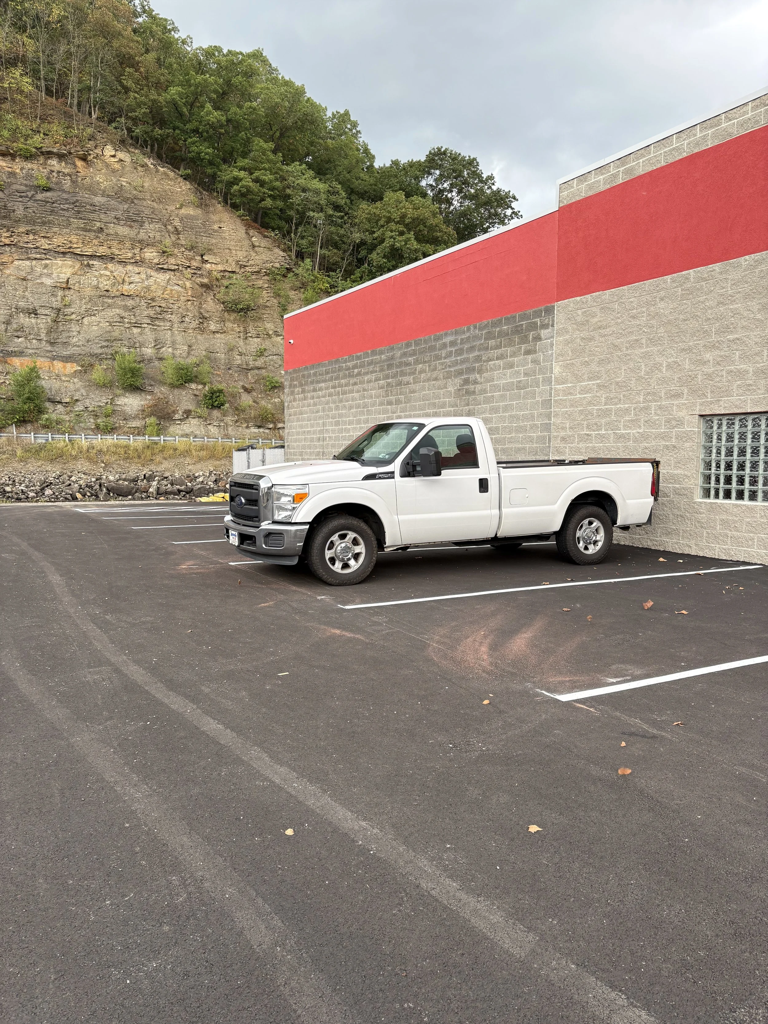 A white pickup truck parked in an empty parking lot next to a building with a red stripe on the wall, with a hillside covered in trees in the background.