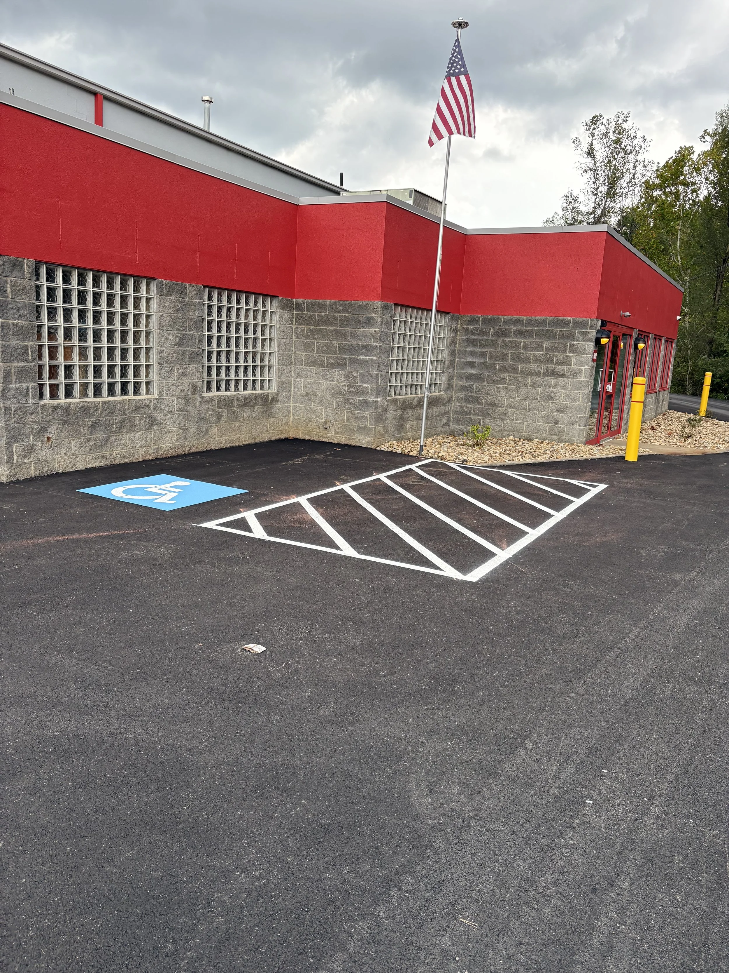 An empty parking space with a handicapped accessible sign next to it, in front of a red building with an American flag flying on a pole. The parking lot has fresh black asphalt and white painted lines.