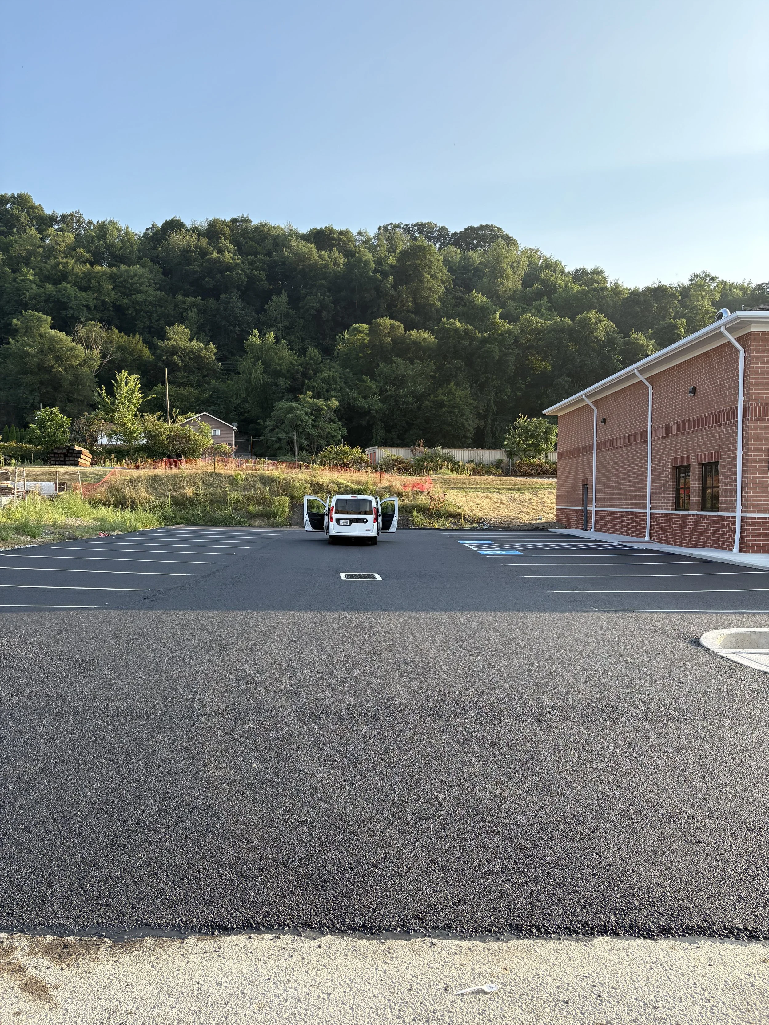Empty parking lot with a white van parked, a building on the right, green trees and hillside in the background, and a clear blue sky.