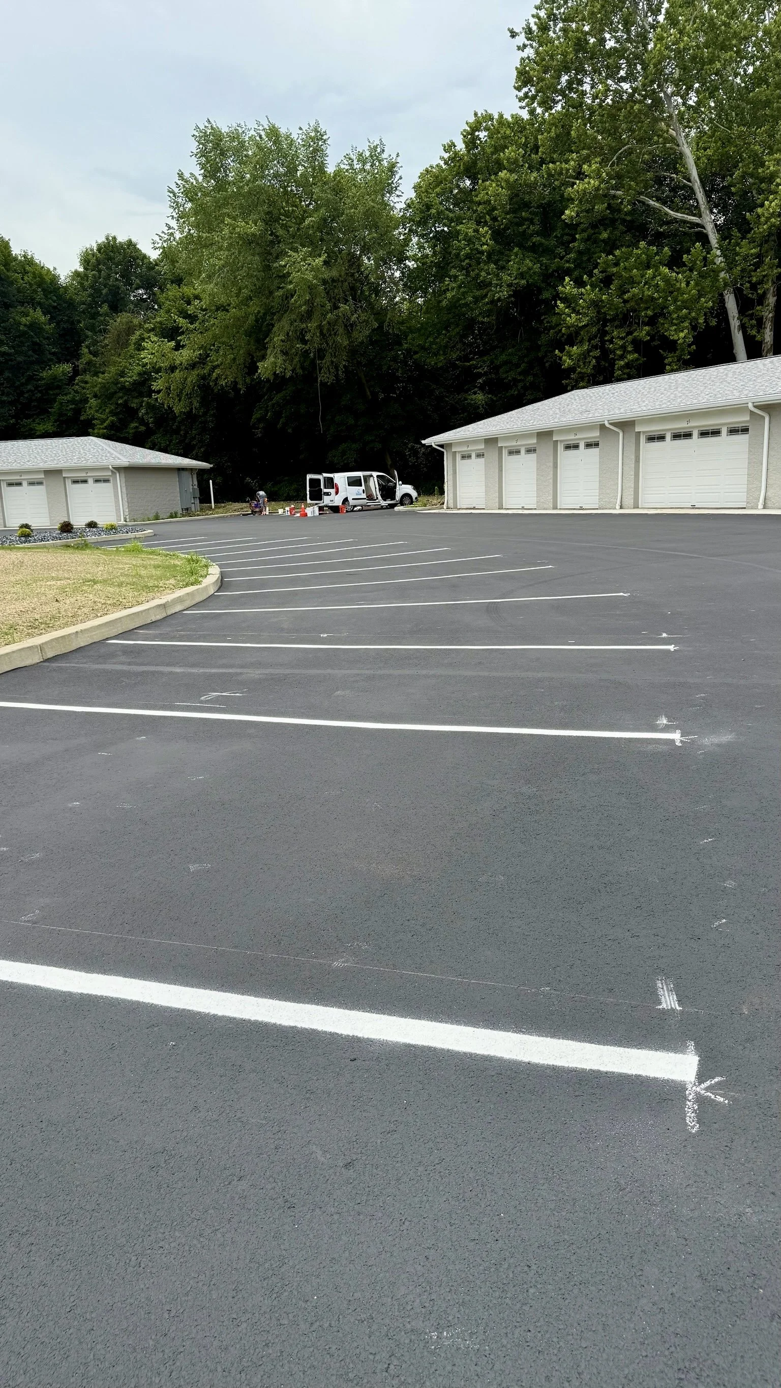 Empty parking lot with white parking space lines, two white garages, and a wooded area in the background.