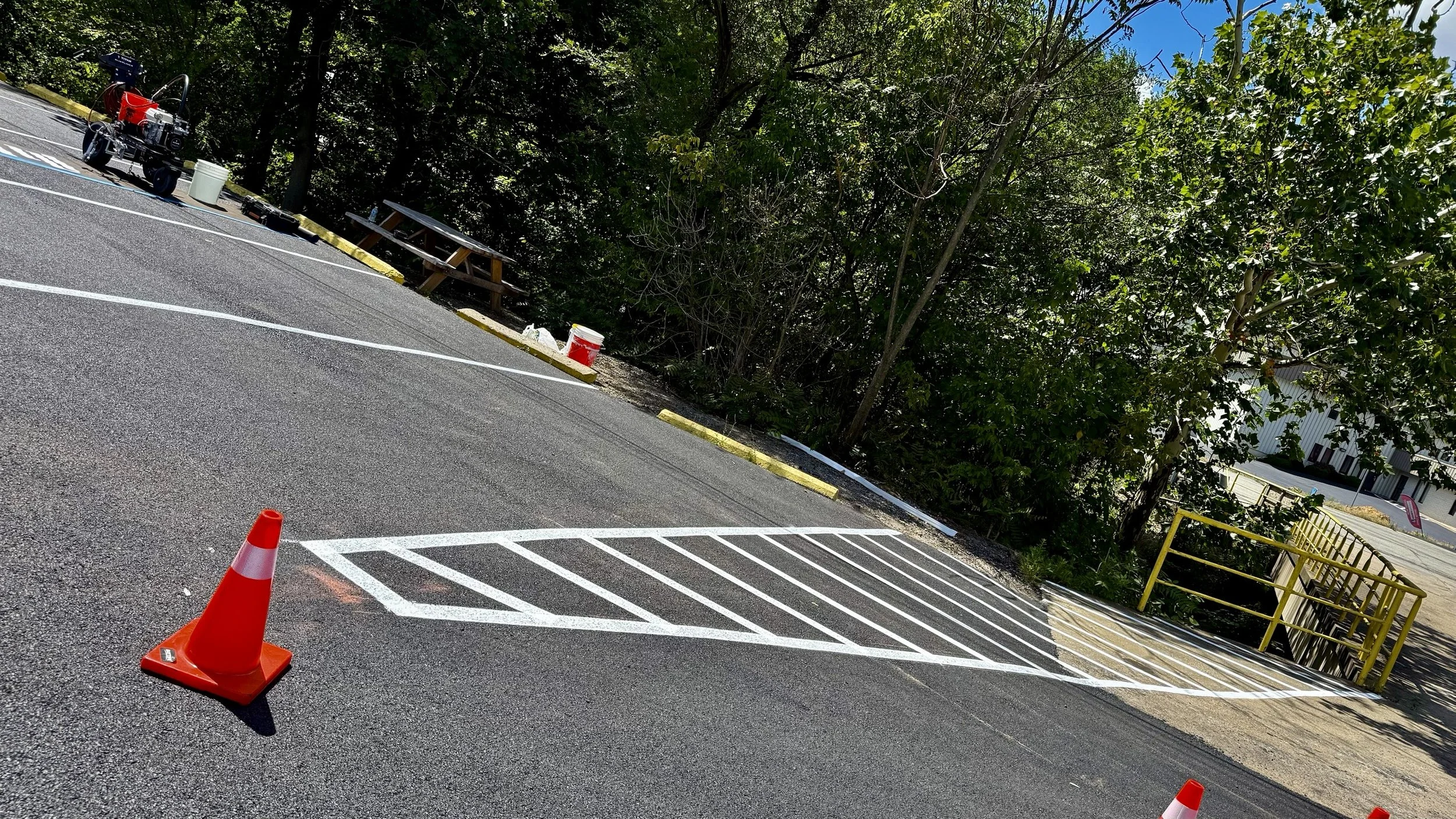 A parking lot section with white painted lines and orange traffic cones, backed by trees and a building in the background.