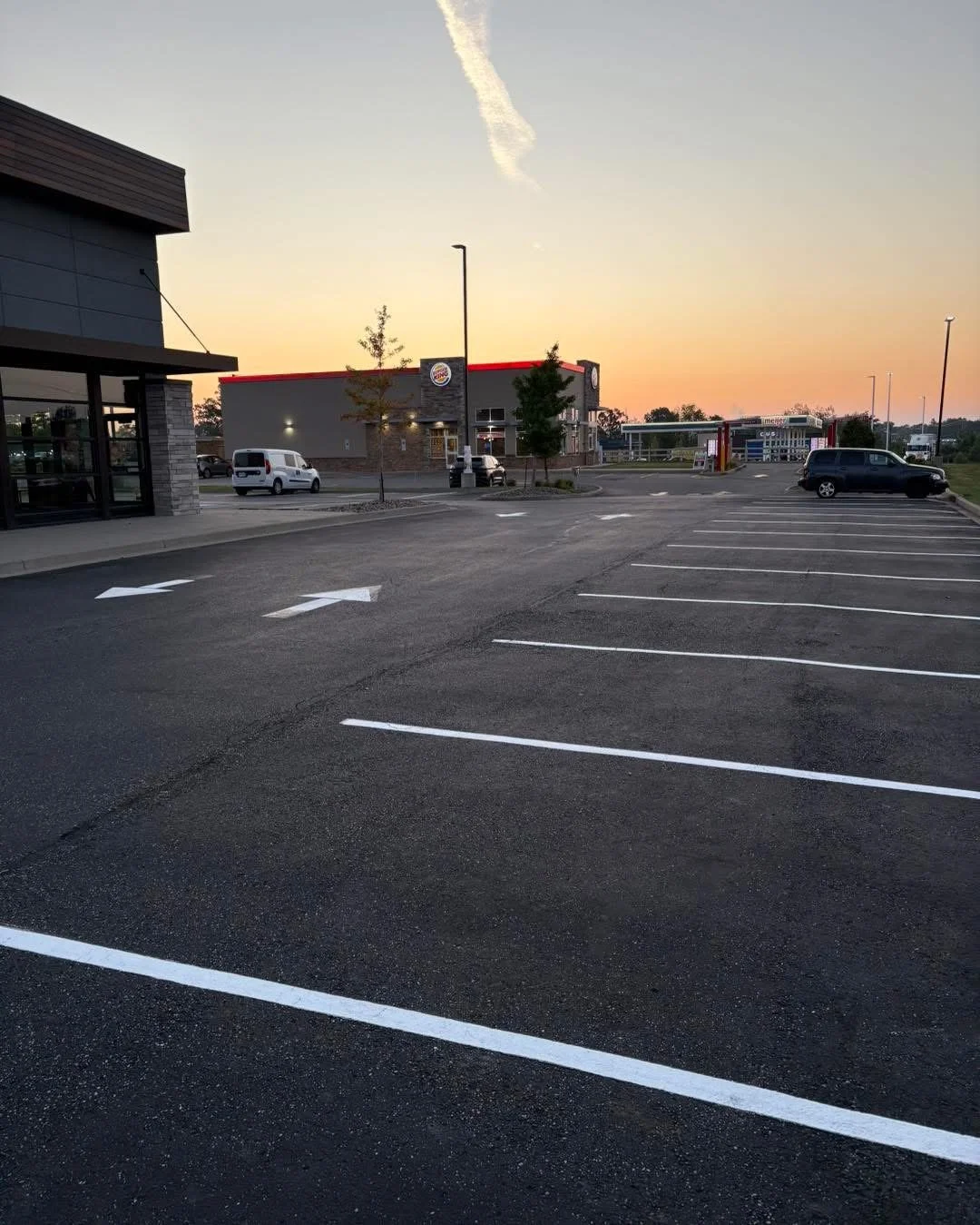 An empty parking lot outside a fast food restaurant during sunset with a few parked cars and clear sky.