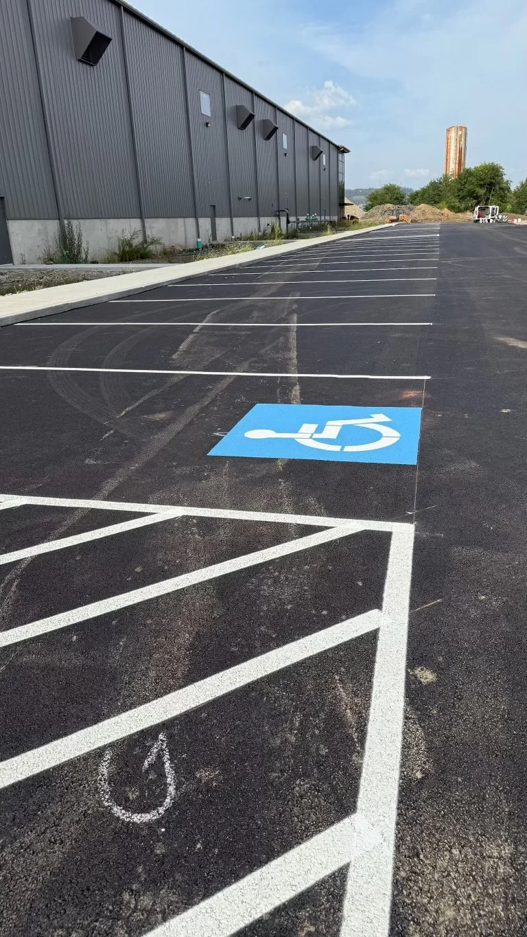 Empty parking lot with handicap accessibility parking space and lines for other parking spots next to a grey industrial building under a partly cloudy sky.