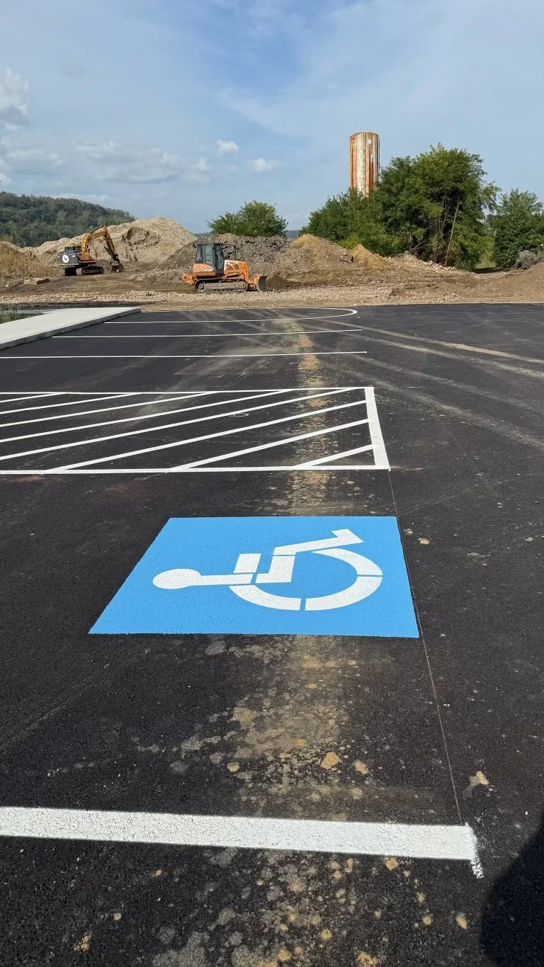 Parking lot with a dedicated handicapped parking space marked by a blue symbol of a person in a wheelchair, with construction equipment and dirt piles in the background under a partly cloudy sky.