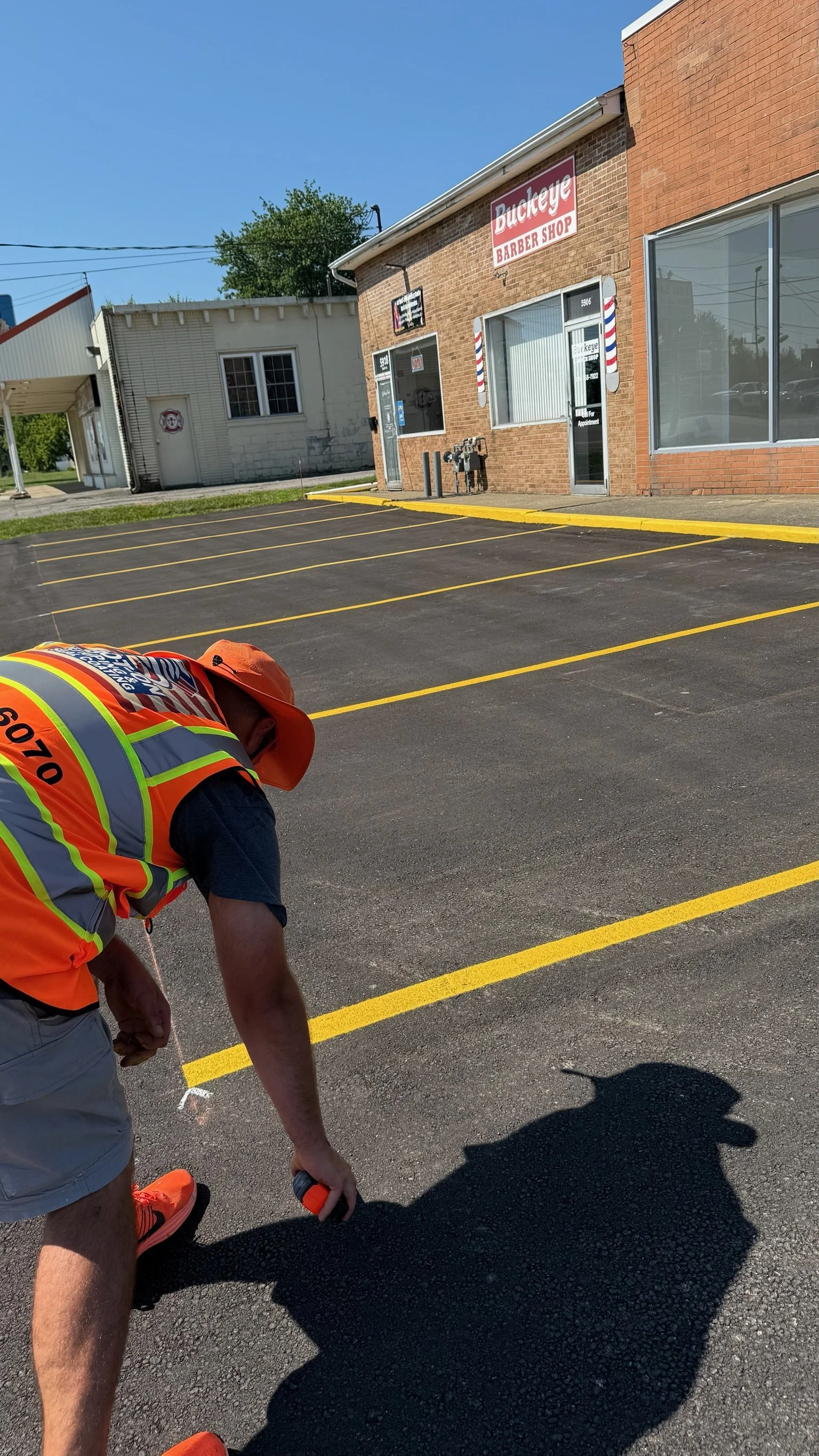 A man in a reflective vest and hat is working in a recently paved parking lot outside a building labeled 'Ducky' Barbershop, with yellow parking lines and a clear blue sky.