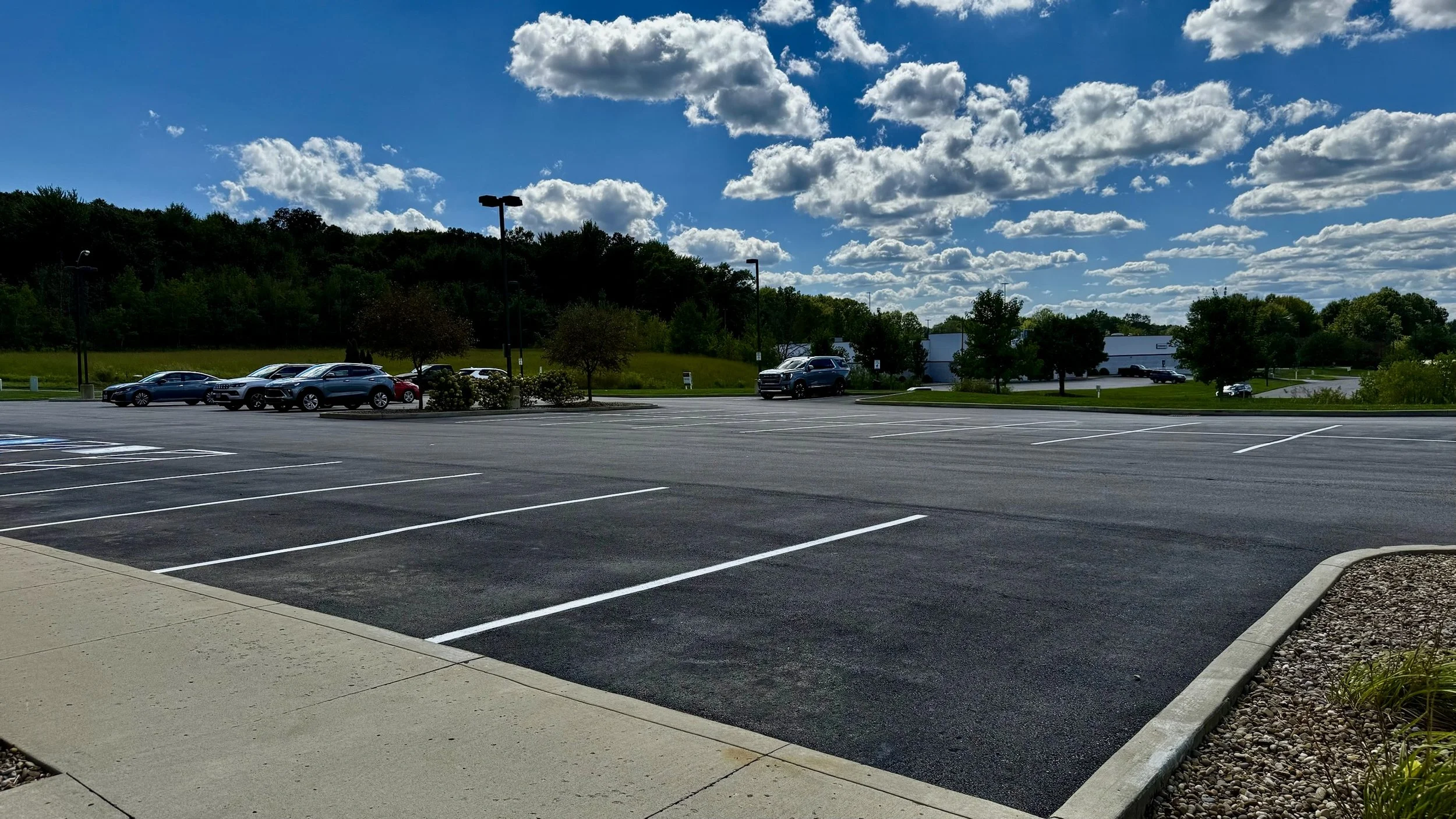 Empty parking lot under blue sky with scattered clouds, few cars parked along the edge, in a suburban area with trees and buildings in the background.