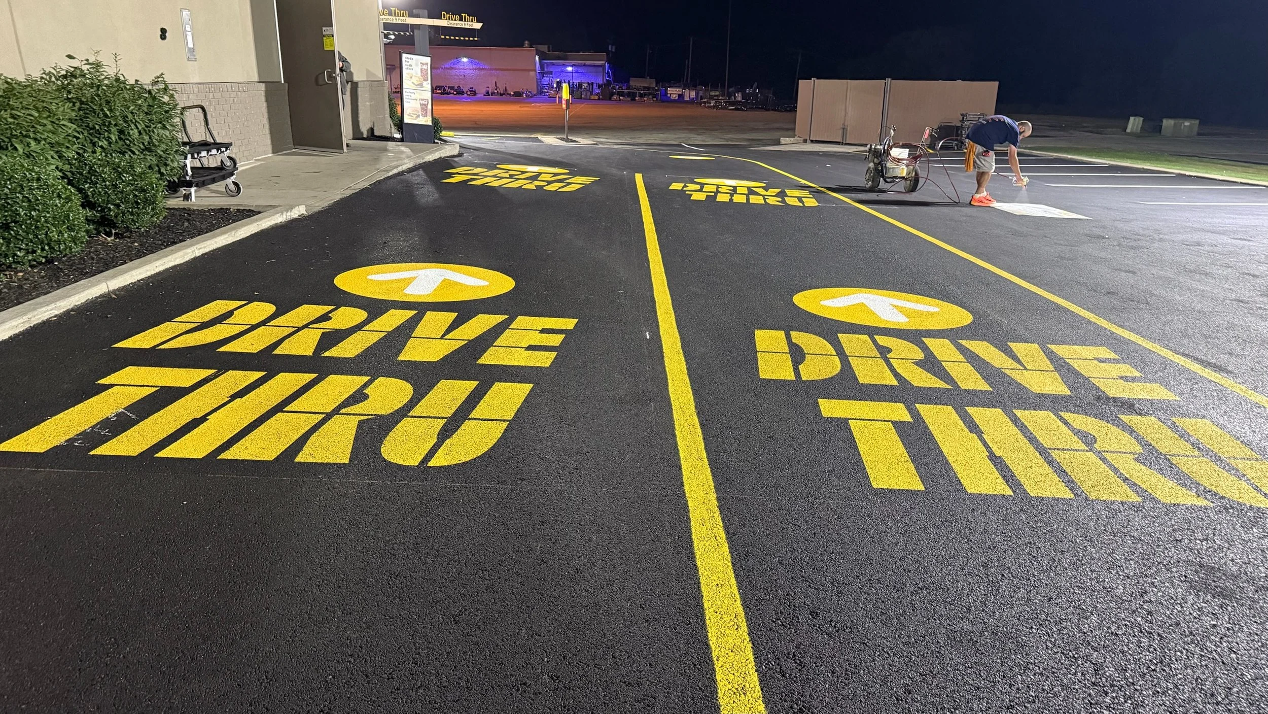 Nighttime view of freshly painted yellow parking lot signs on asphalt, instructing 'Drive Thru' with arrows, and 'Drive' with arrows, in front of a building, with a person painting a crosswalk in the background.