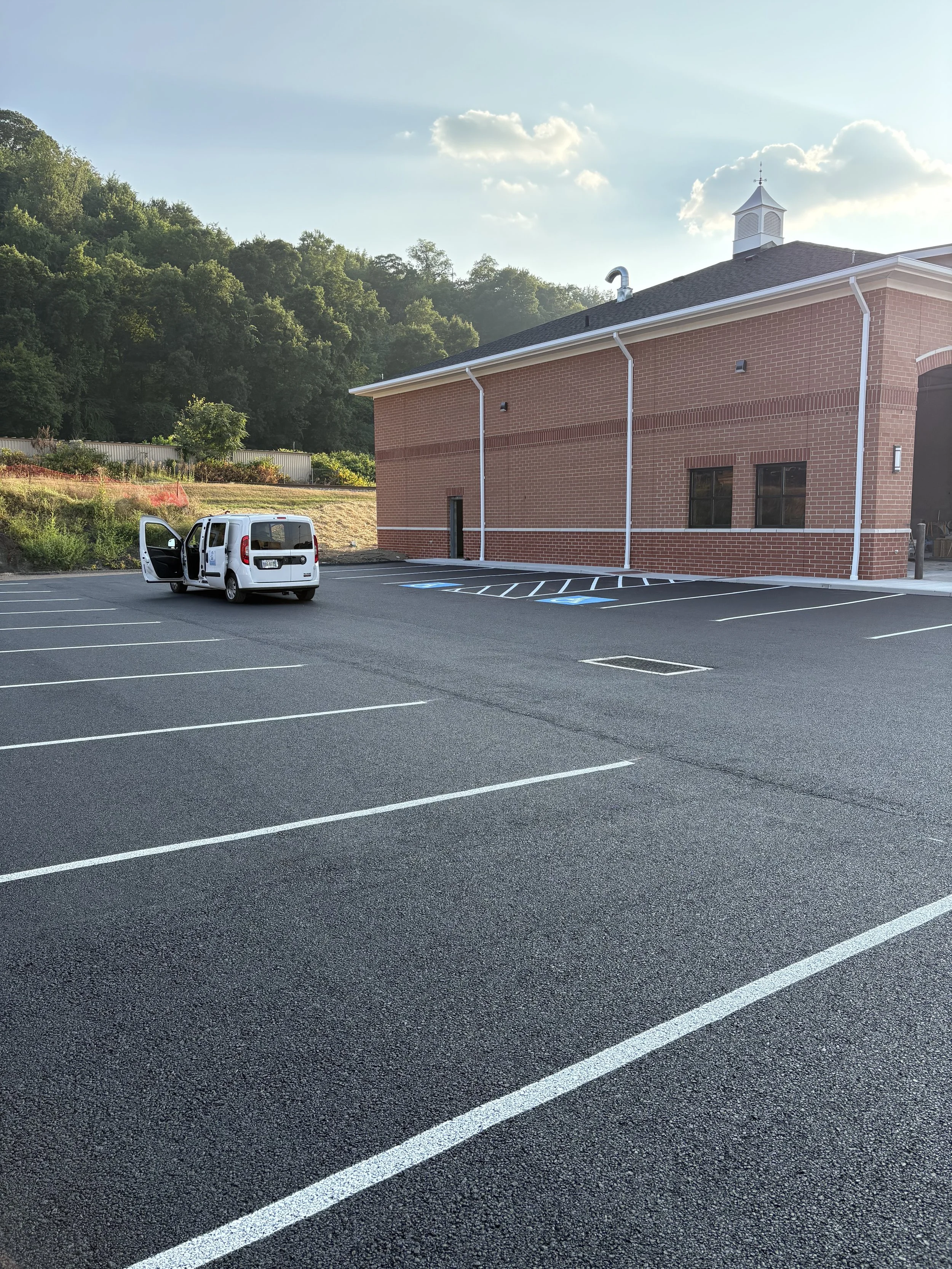 Empty parking lot with a white van and an open door, a brick building, and a hill with trees in the background, under a partly cloudy sky.