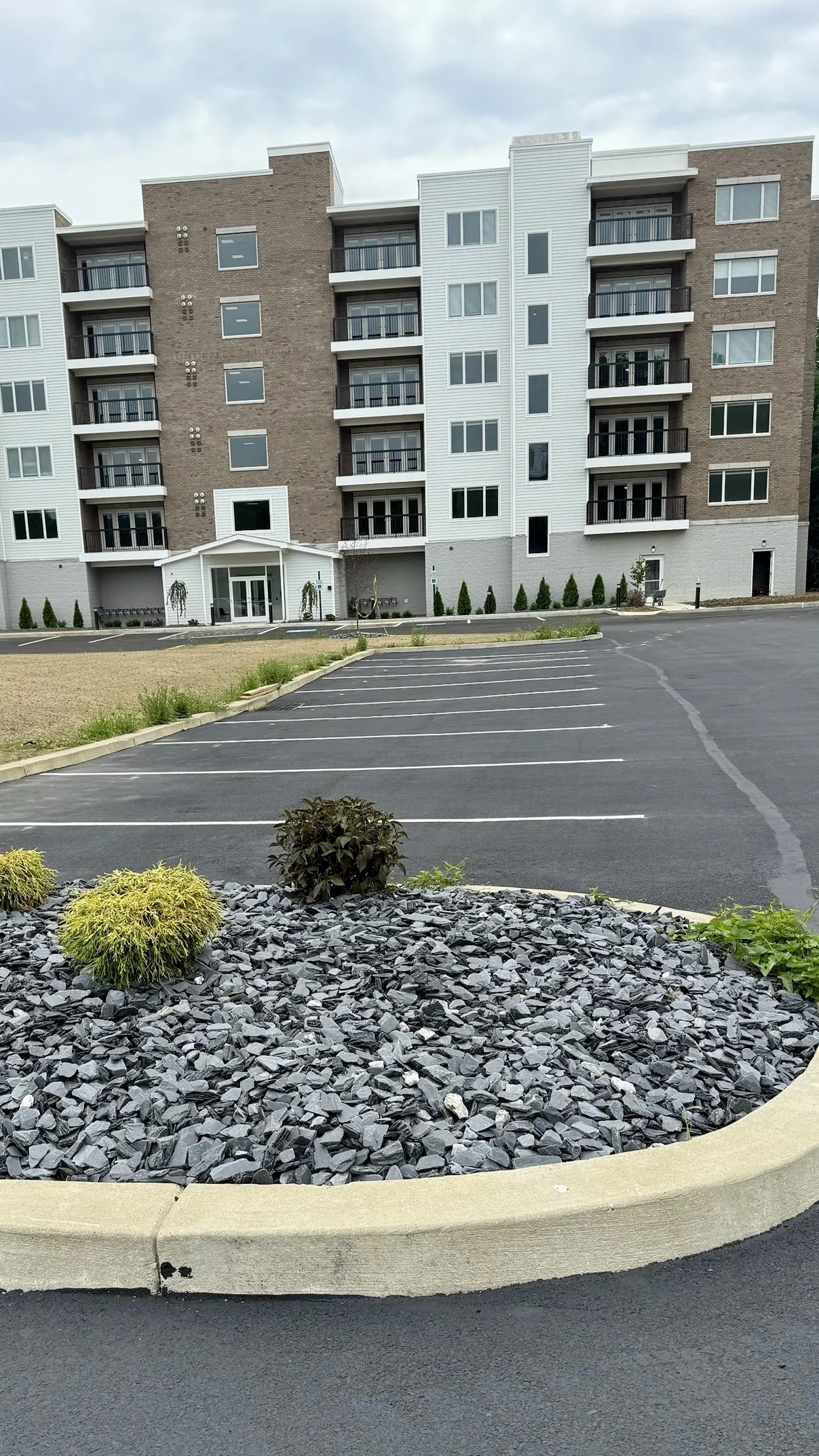Exterior view of a modern multi-story apartment building with balconies, surrounded by a paved parking lot and landscaped area with bushes and rocks.