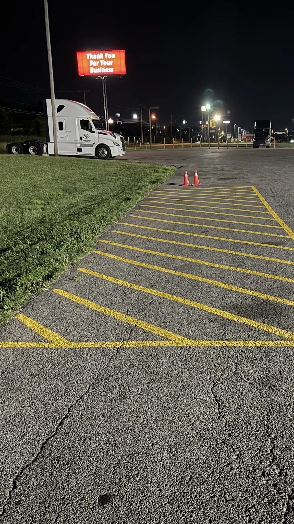 Empty parking lot with yellow striped area, two orange traffic cones, a white semi-truck, and a digital sign reading "Thank You For Your Business" at night.