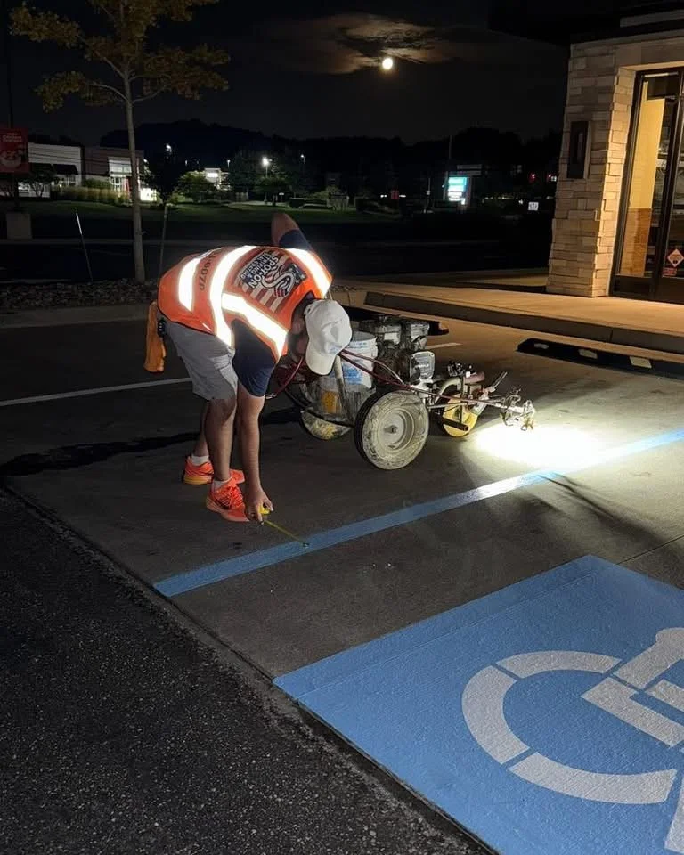 Construction worker painting a handicap parking space blue at night with a bright light, using a laser level, outside a building under a full moon.