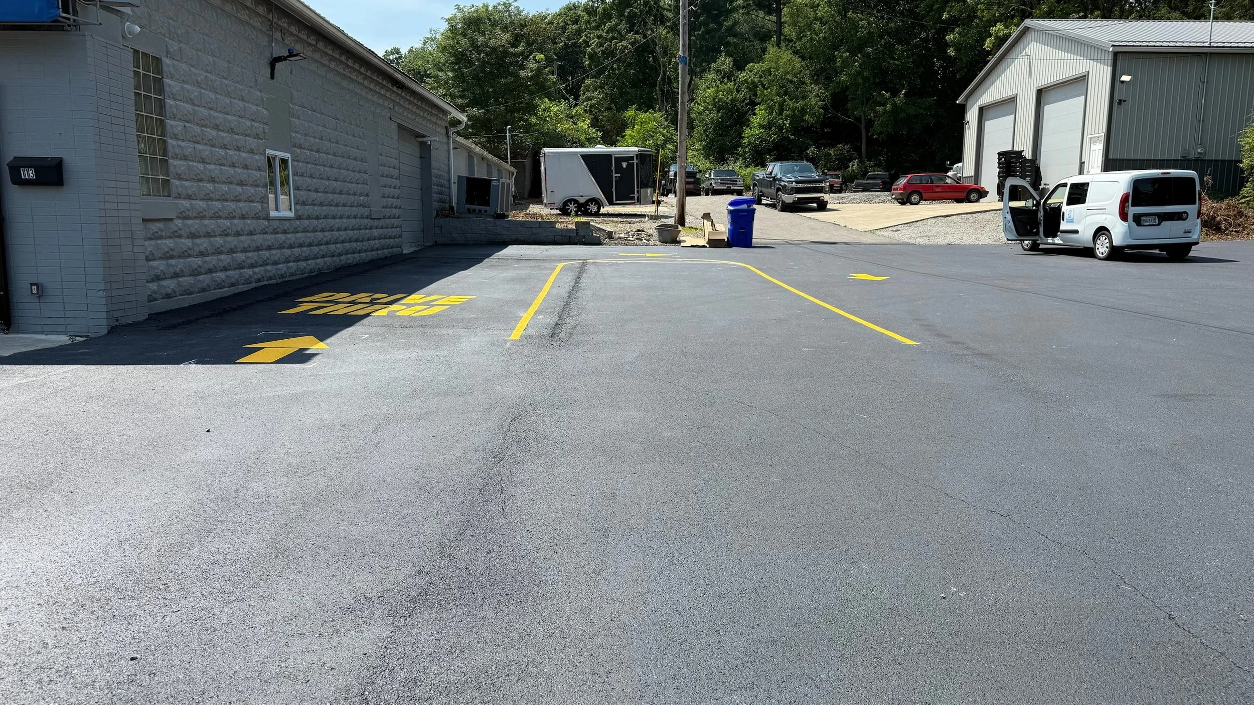 Empty parking lot with freshly painted yellow parking space lines and directional arrows. A white van with an open door is parked on the right, with other vehicles and buildings visible in the background.