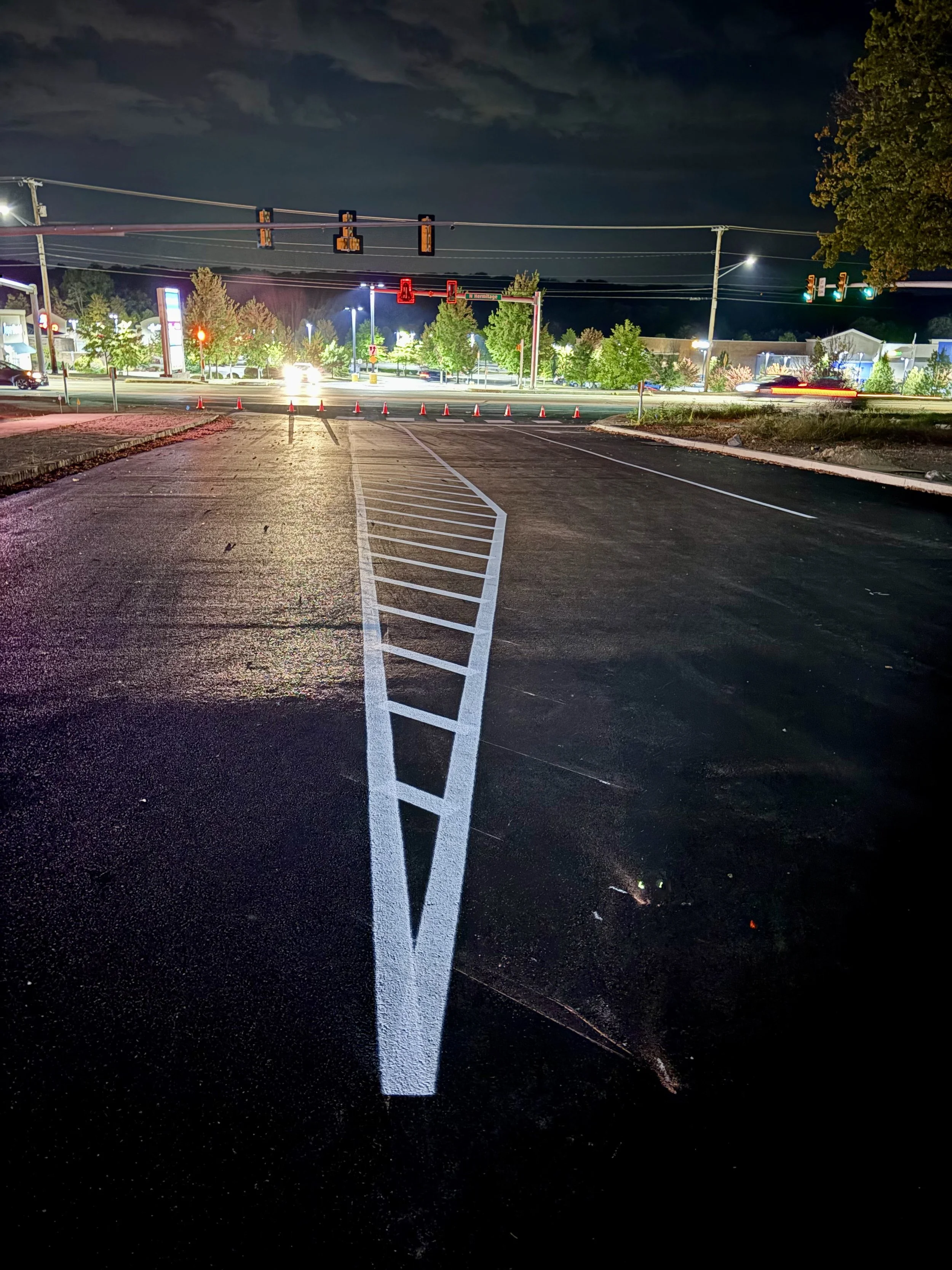 Empty asphalt parking lot at night with white painted lines and cones blocking the road, traffic lights and illuminated stores in the background.