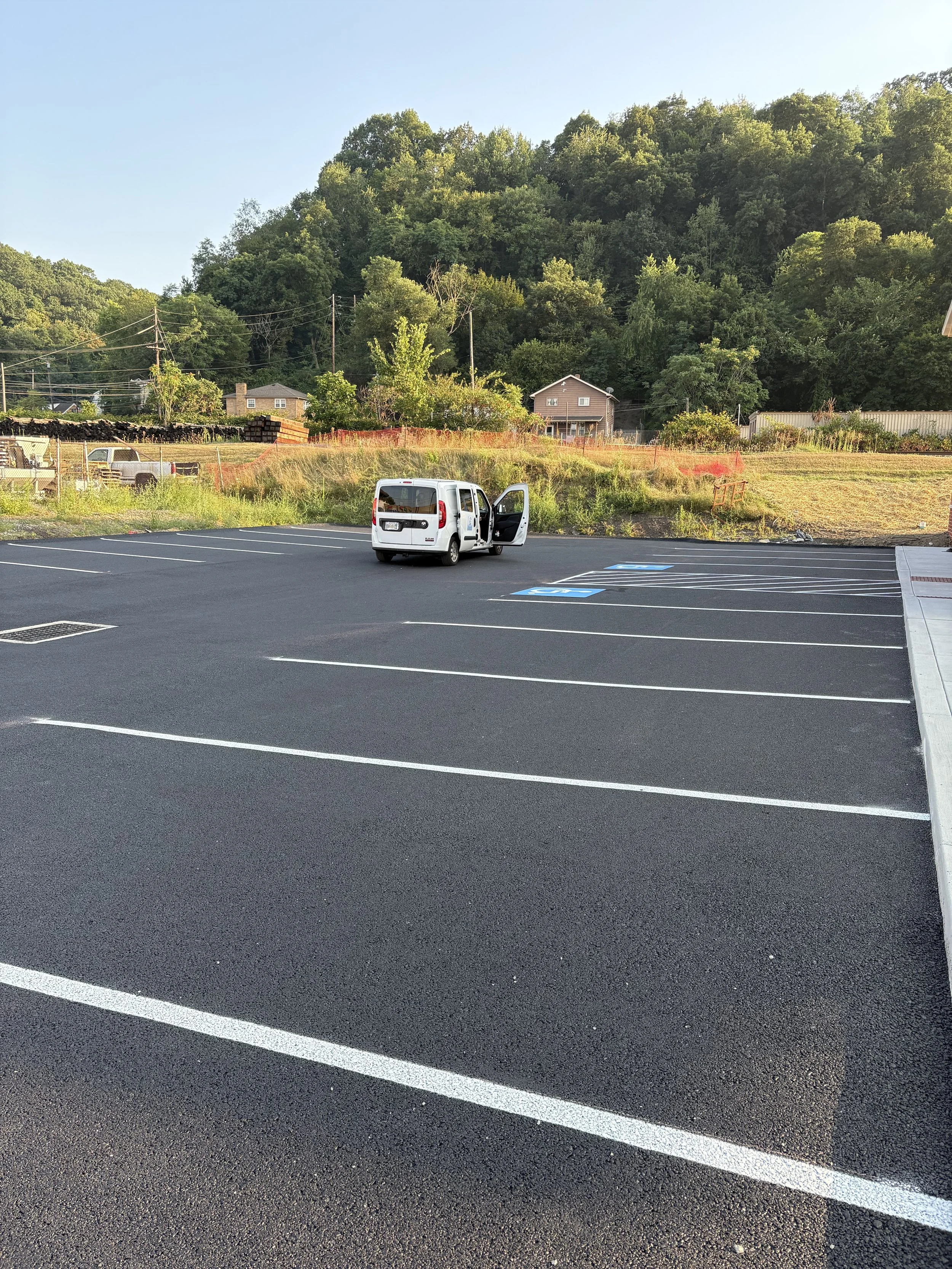 Empty parking lot with a white van parked and first parking space occupied. Background shows a hillside with trees, houses, and a partly cloudy sky.