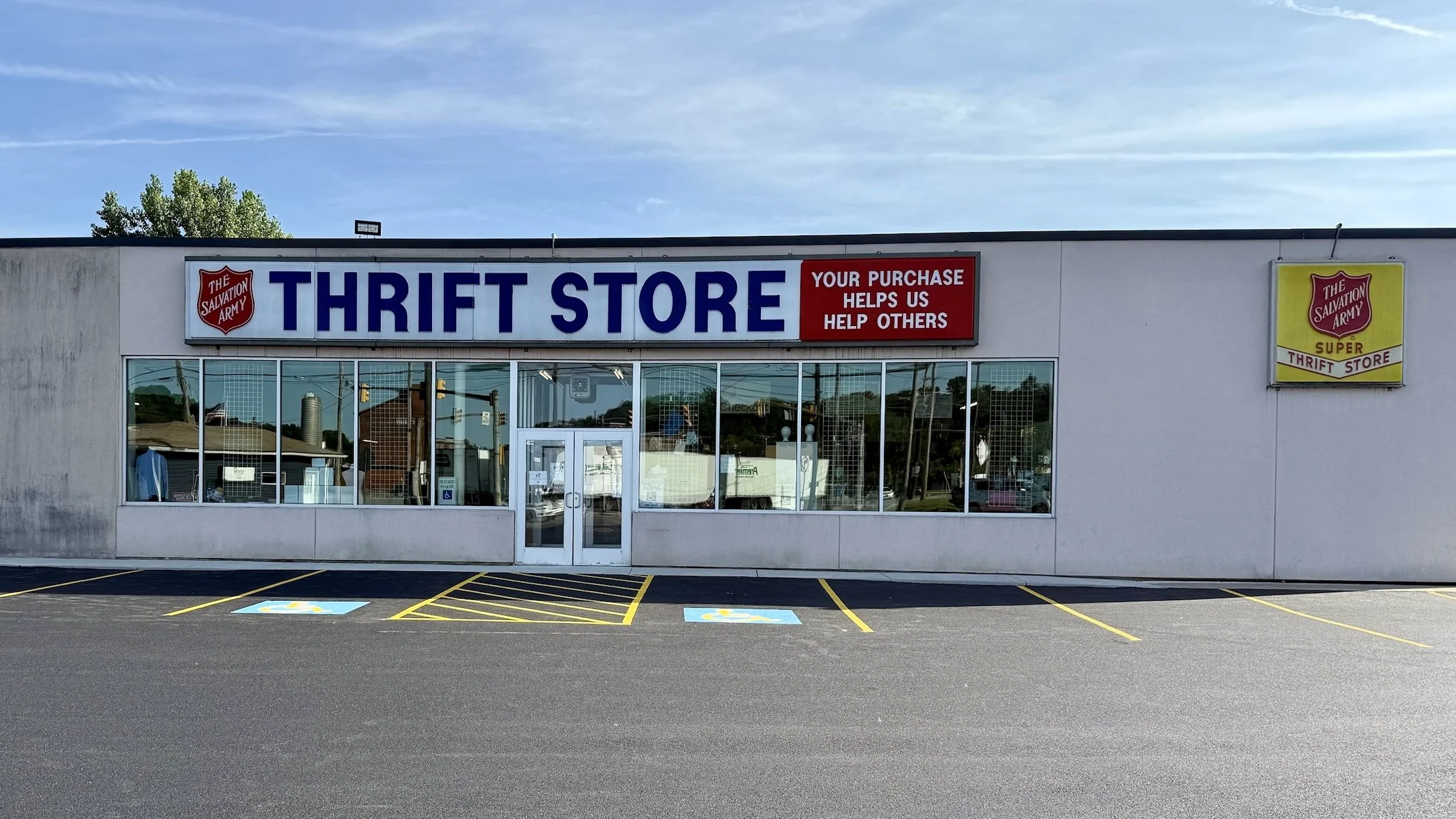 Front view of a Salvation Army thrift store with a large sign that reads 'THRIFT STORE' and a red banner stating 'Your purchase helps us help others.' There are two parking spaces marked for handicapped parking in front.