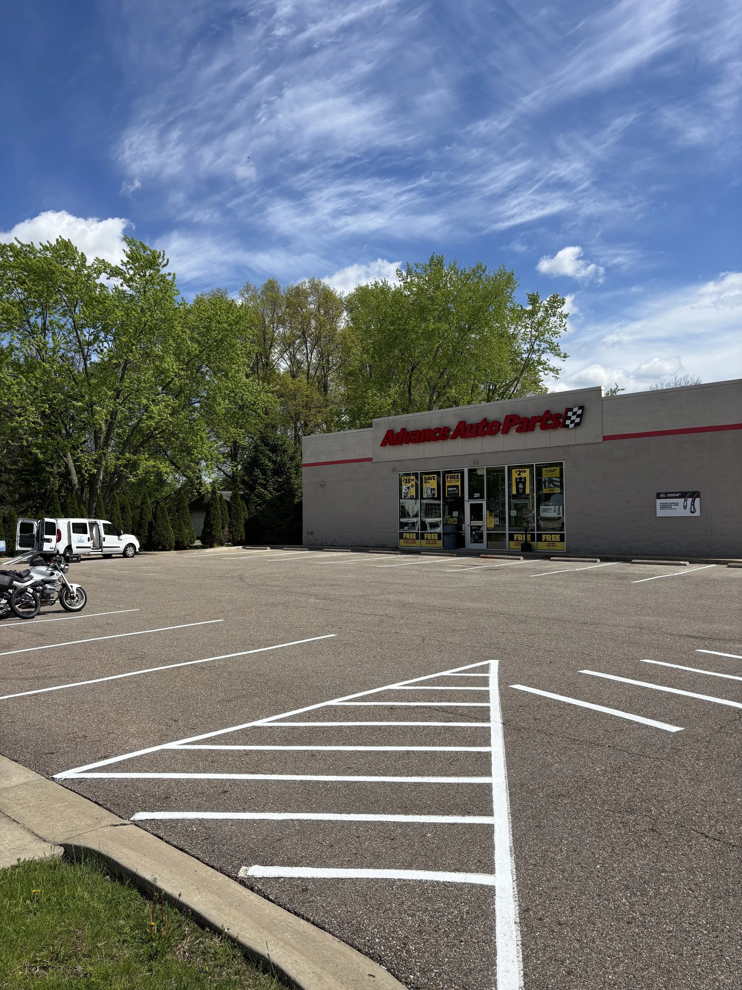 Empty parking lot in front of an Advance Auto Parts store with motorcycles and vans parked, green trees behind the store, and a blue sky with clouds overhead.