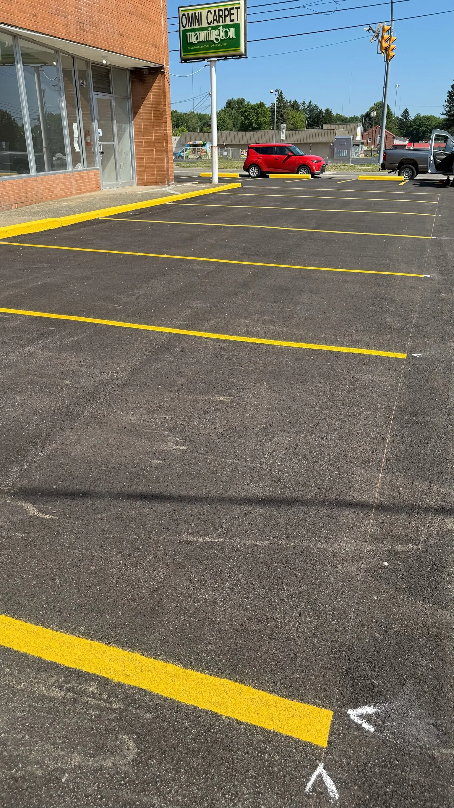Empty asphalt parking lot with yellow painted parking spaces and white arrows, next to a brick building with glass windows and a sign for OMNI CARPET, with a red car parked in the background under a clear blue sky.