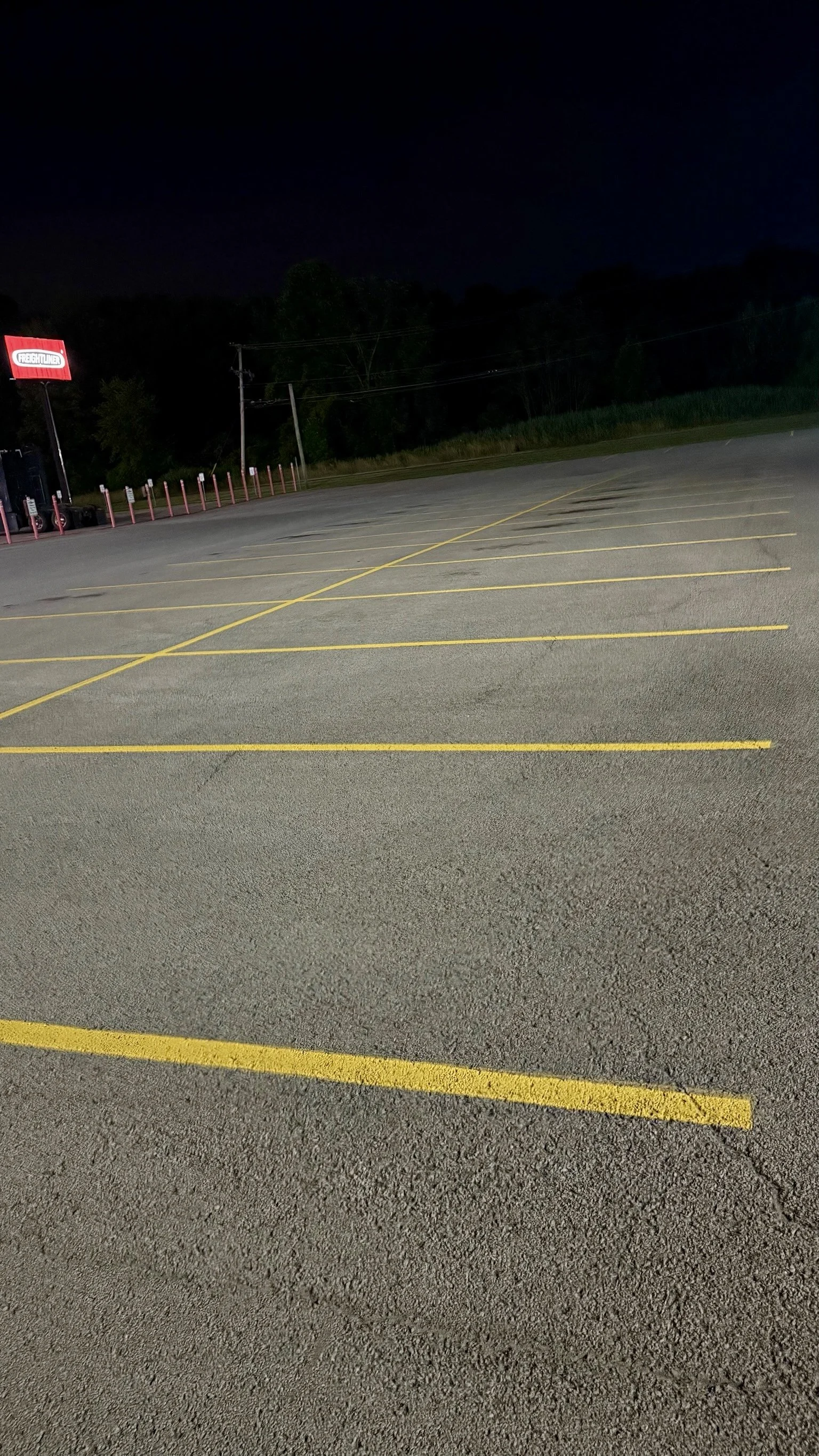 Empty parking lot at night with yellow parking space lines and a red sign for Freightliner in the distance.