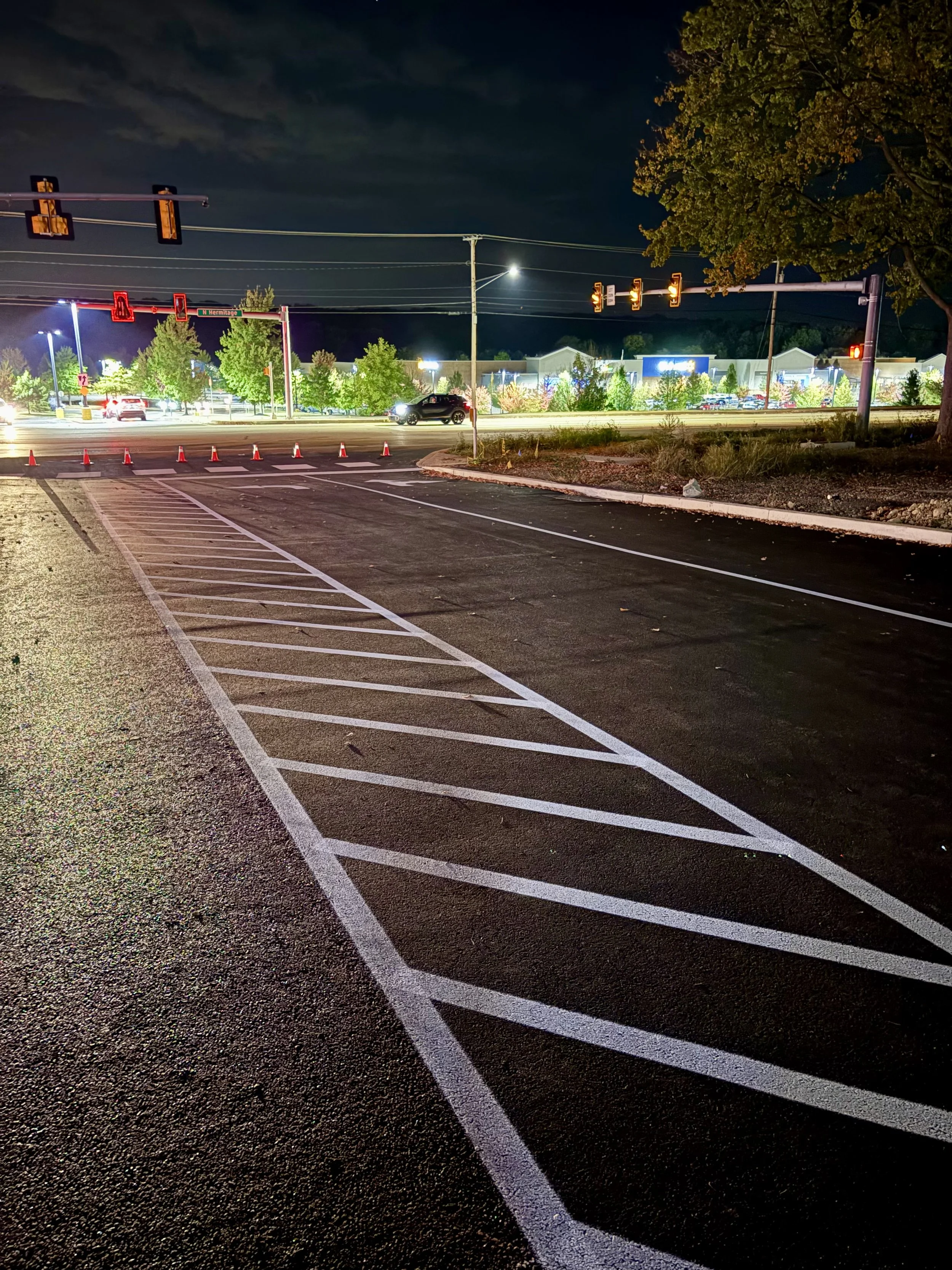 Nighttime view of a parking lot with empty diagonal striped spaces in the foreground, a busy street with cars, traffic lights, and buildings illuminated in the background.