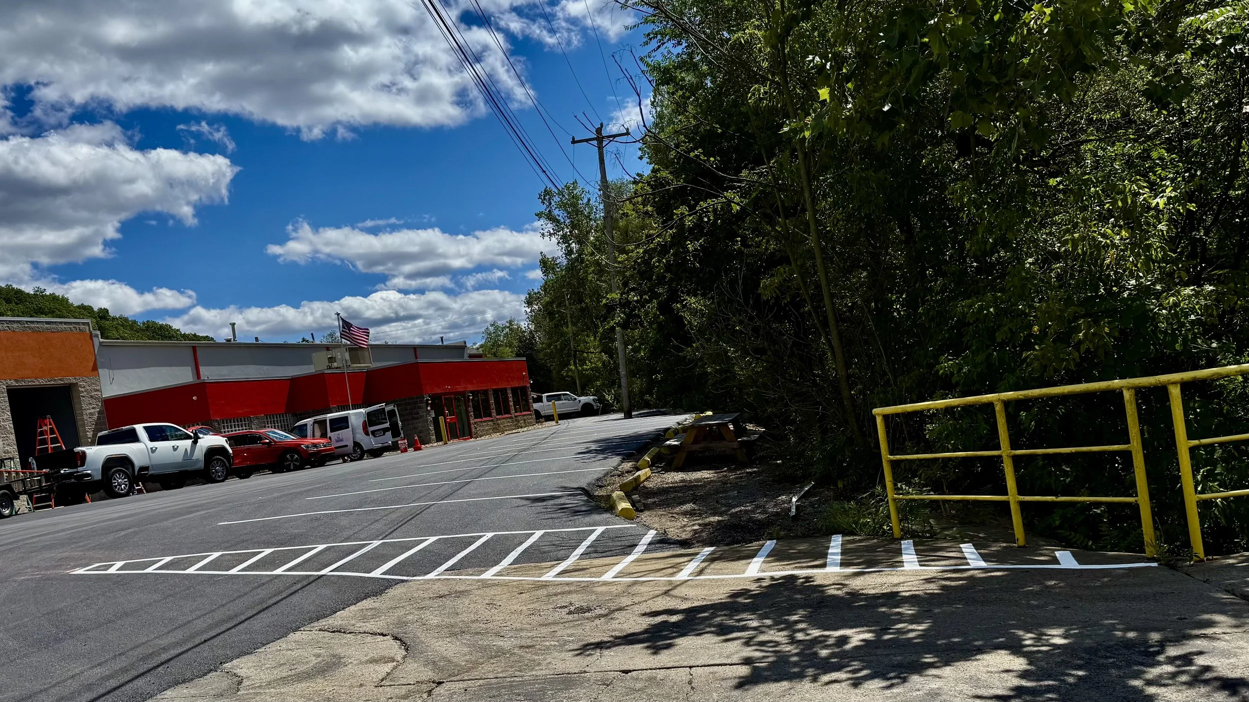 The image shows the exterior of a commercial building, likely a store or restaurant, with red and gray walls. There are several parked vehicles in the parking lot, including pickup trucks and vans. The American flag is flying near the building. On th