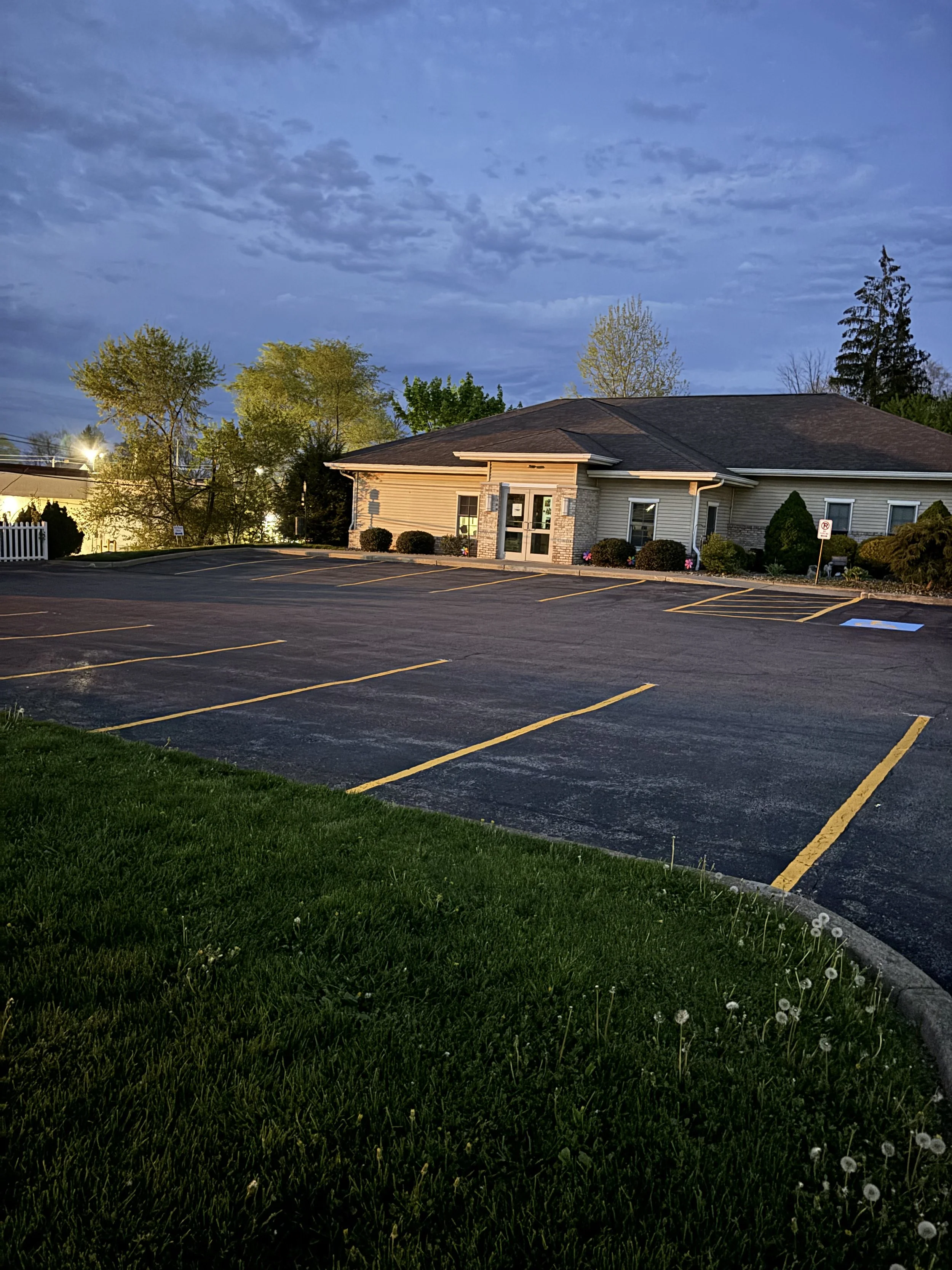 Empty parking lot in front of a commercial building during early evening with some trees and a partly cloudy sky in the background.