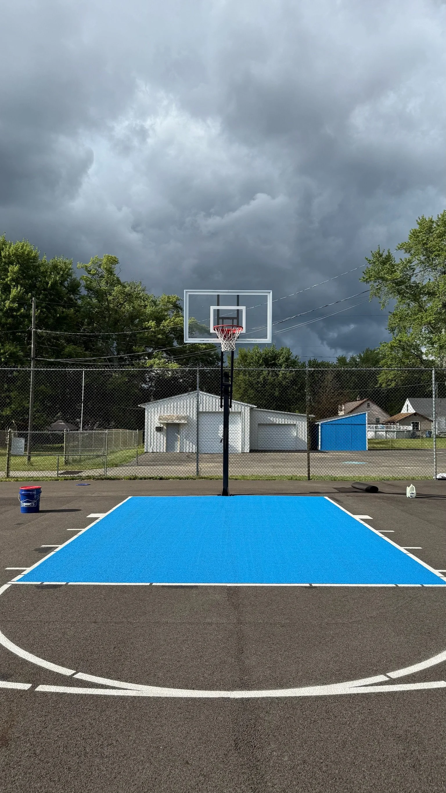 An outdoor basketball court with a blue painted section in front of the hoop, surrounded by a chain-link fence. The sky is overcast with dark clouds, and there are trees and small buildings in the background.