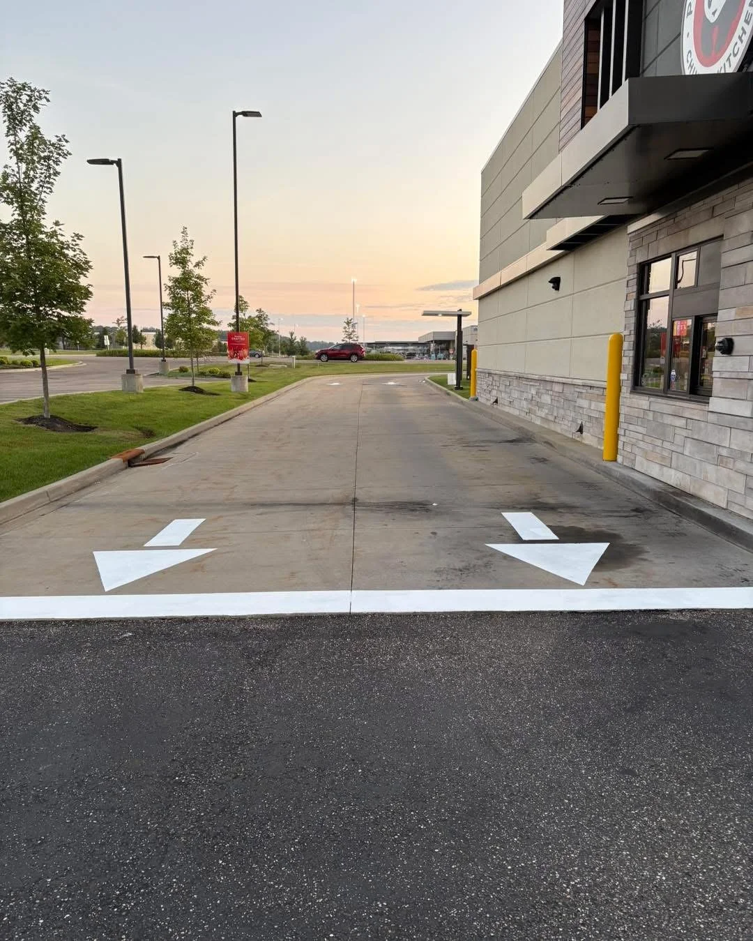 Empty drive-thru lane with white arrows pointing left and right, outside of a fast food restaurant at sunset.