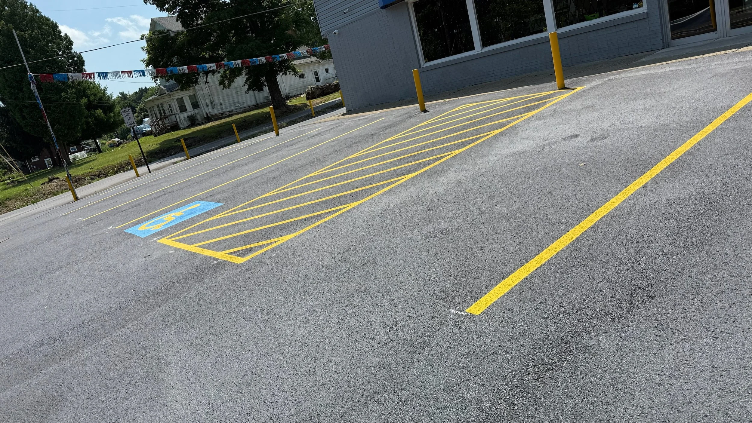 Empty parking lot with designated handicapped parking space marked in blue and yellow, surrounded by yellow posts, in front of a grey building with windows, on a sunny day with trees and houses in the background.