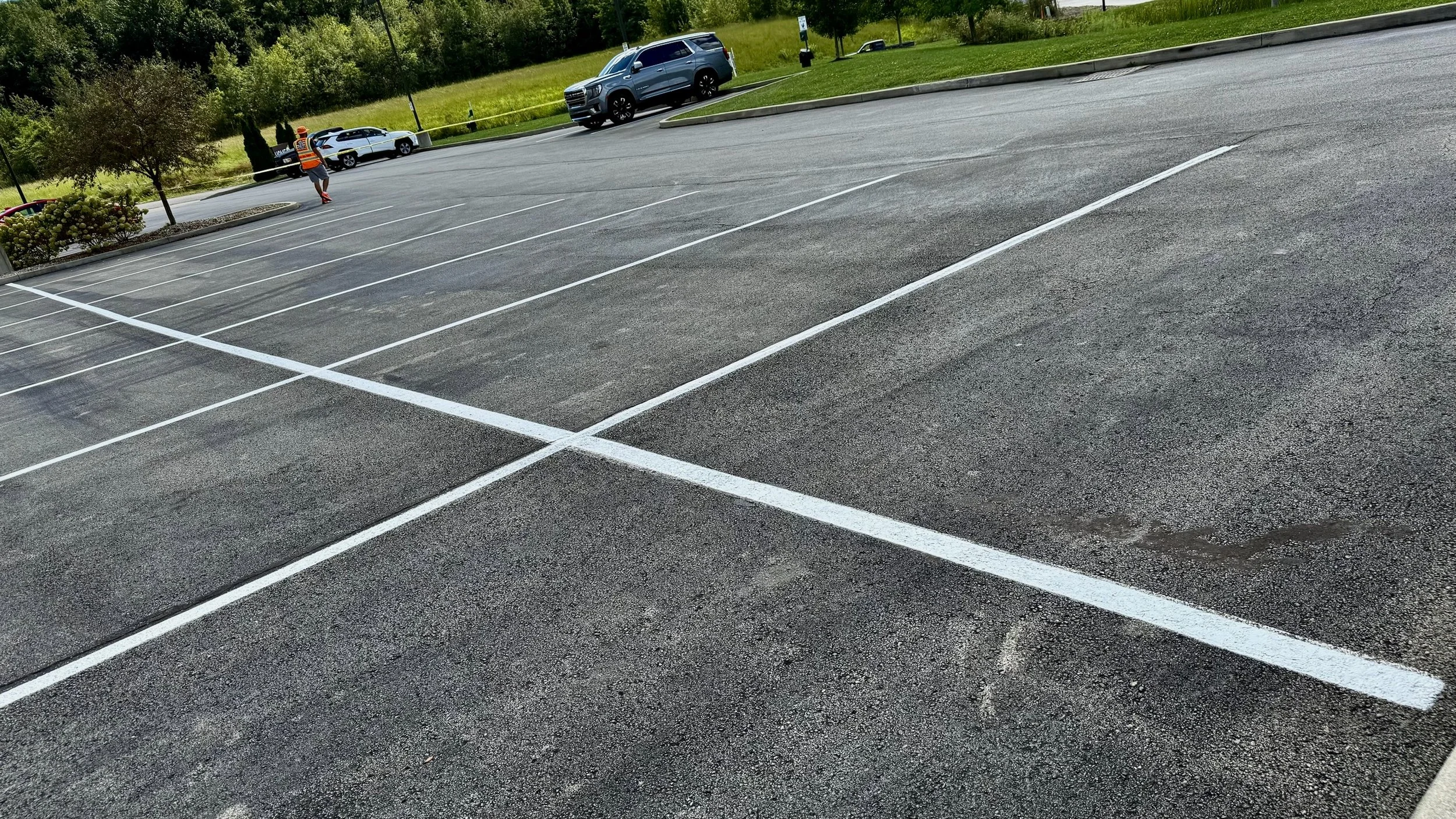 Empty parking lot with white painted parking spaces, a worker in an orange safety vest and helmet, a few cars, trees, and grass in the background.