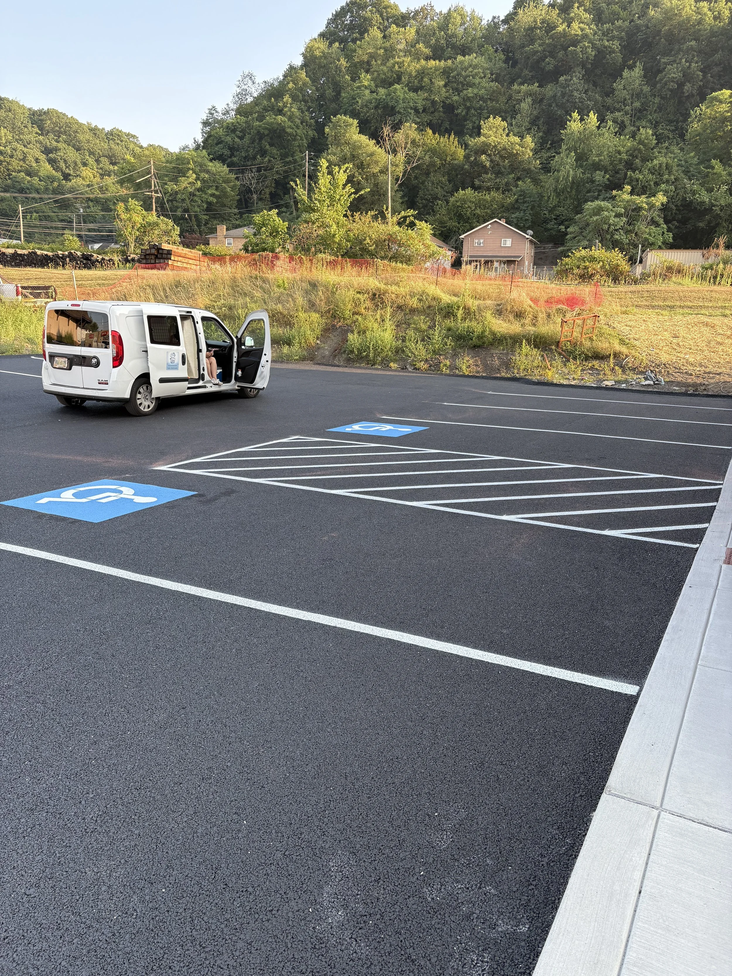 Empty parking lot with designated handicapped parking spaces and a white van parked nearby, with a backdrop of trees, bushes, and a house on a hillside.