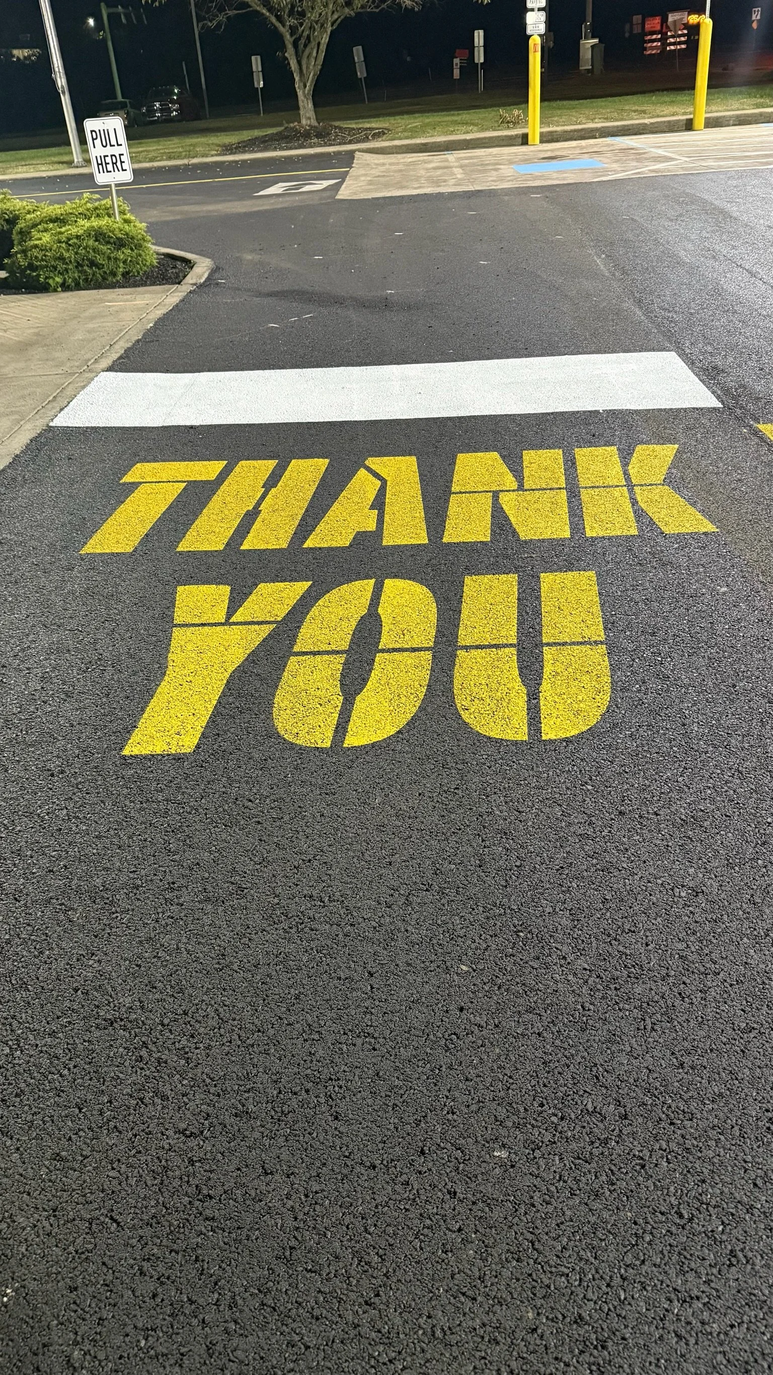 A parking lot surface with yellow painted words saying 'THANK YOU' and a white striped crosswalk. There is a sign on the side that says 'PULL HERE' and a tree in the background.